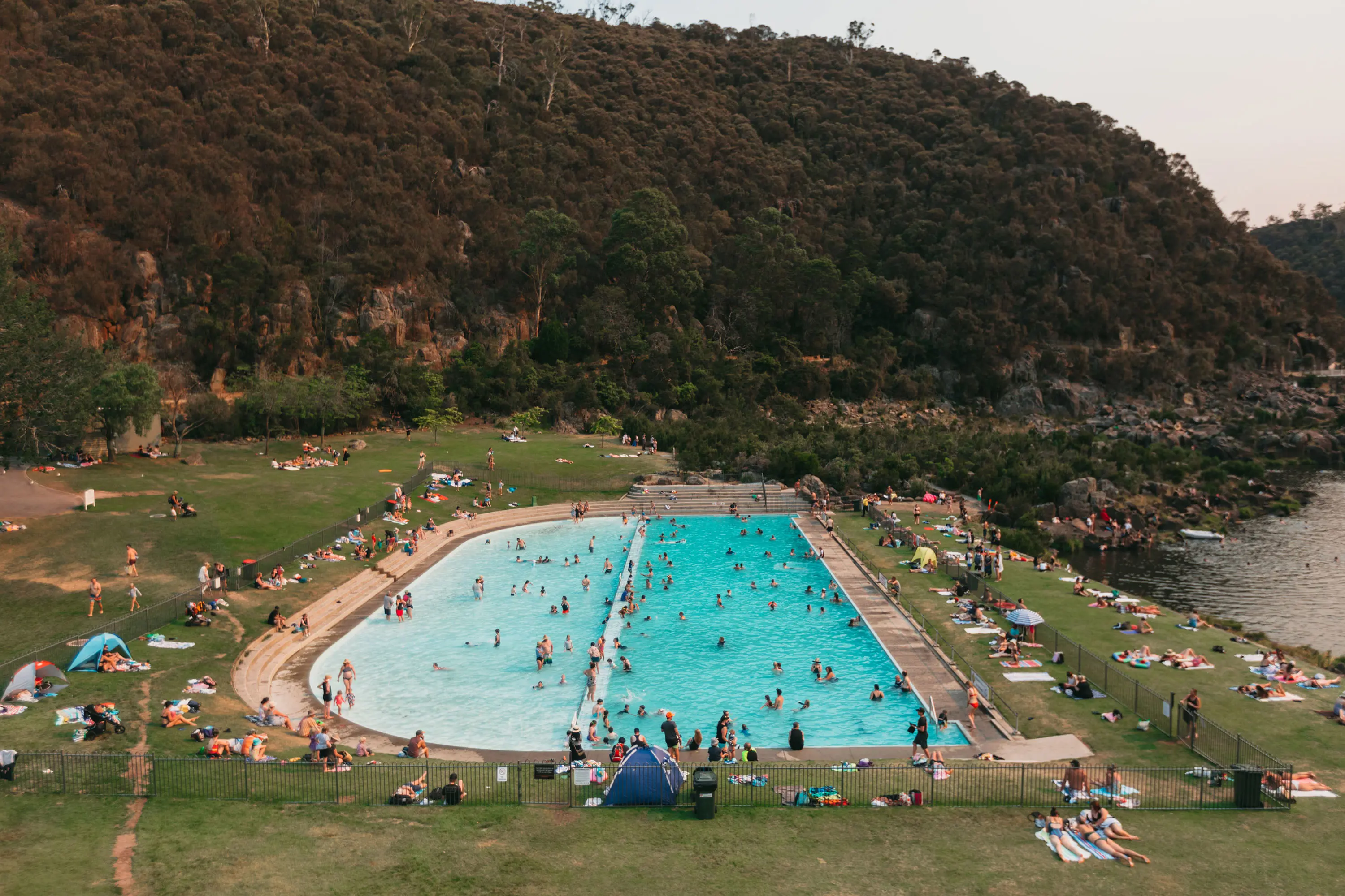 A large, semicircular aqua outdoor pool set into a grassy area next to a river, with lots of people swimming and relaxing. Behind is a steep, densely bushy cliff.