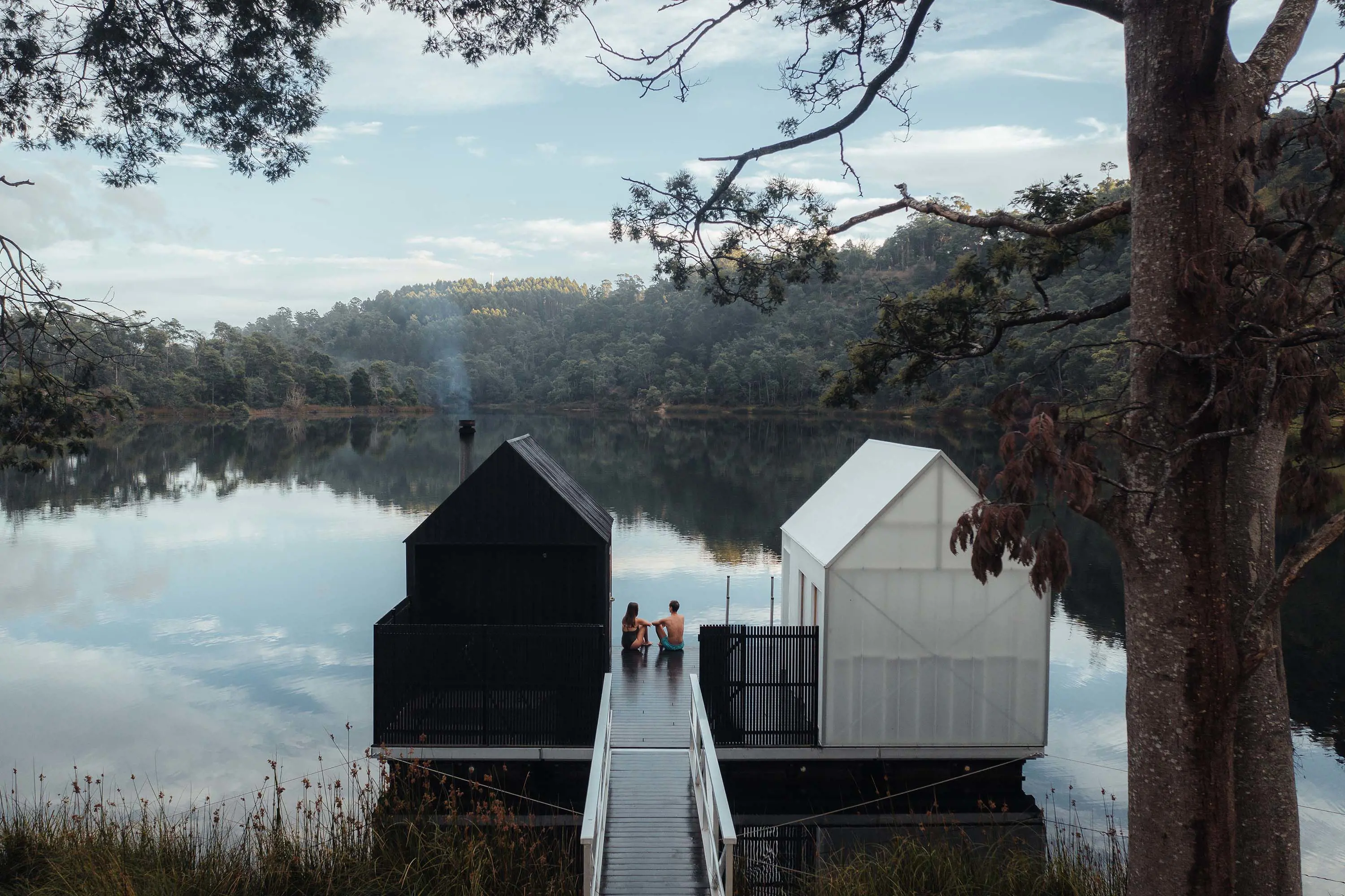 A couple sitting on a wooden jetty between black and white sauna huts, overlooking a lake.