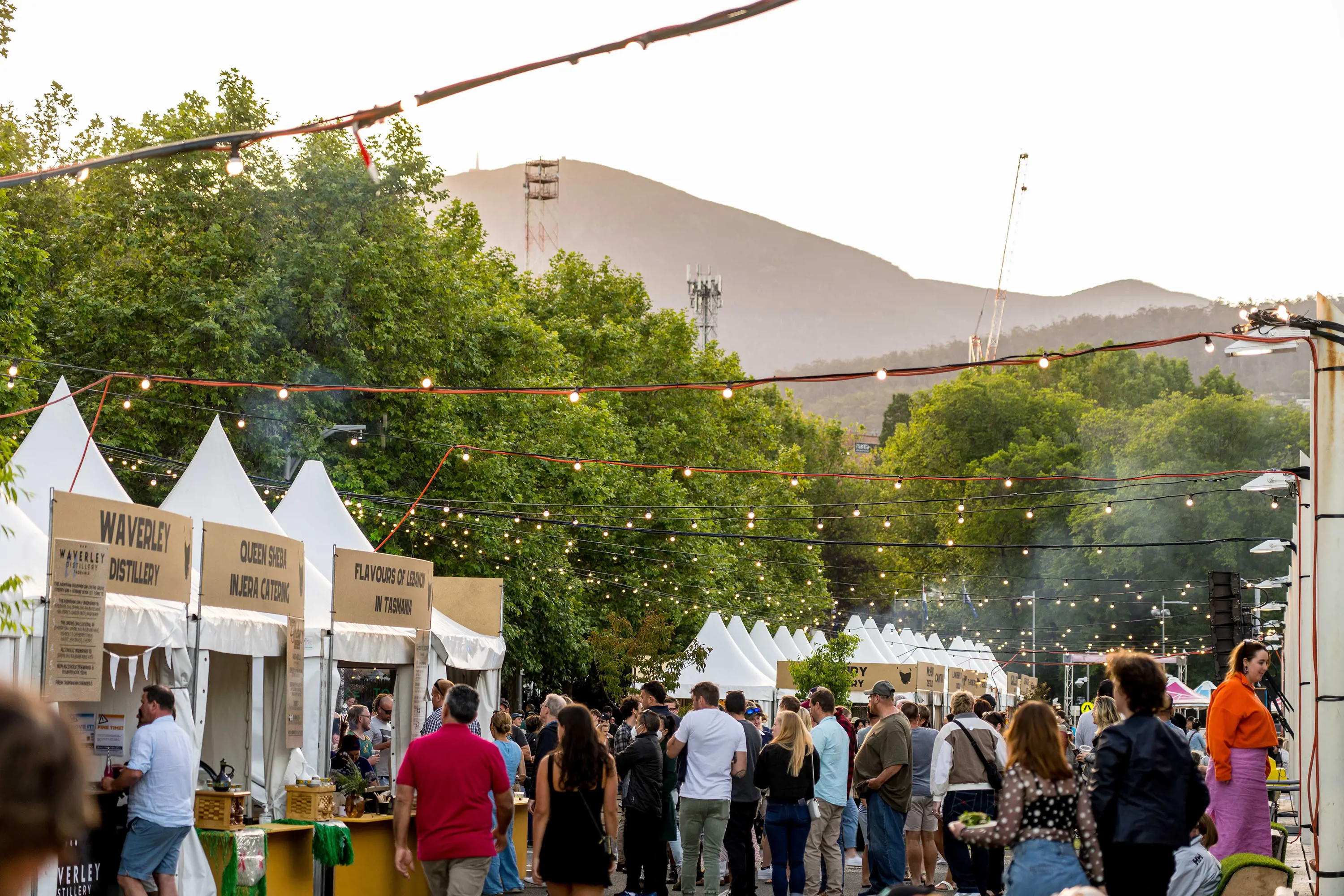 An outdoor market, with white marquee food stalls, crowds of people and strings of lights overhead. In the background, a mountain rises up behind the trees.