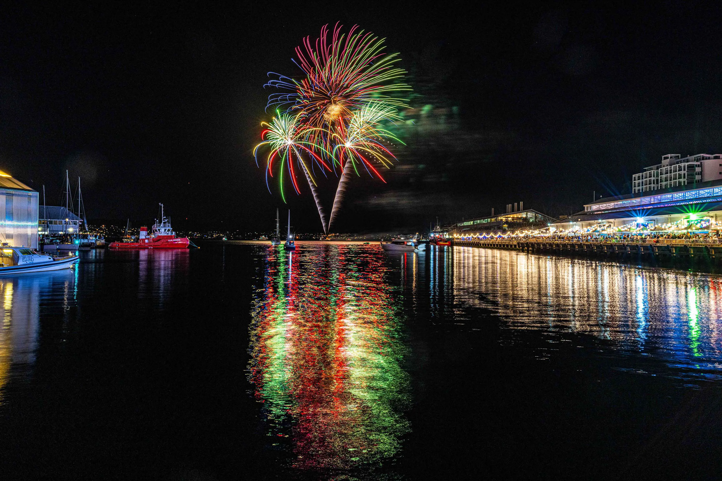 Fireworks shoot up into the sky and are reflected in the dark water of a harbour. Lights from a few boats and buildings on the wharf are reflected also.