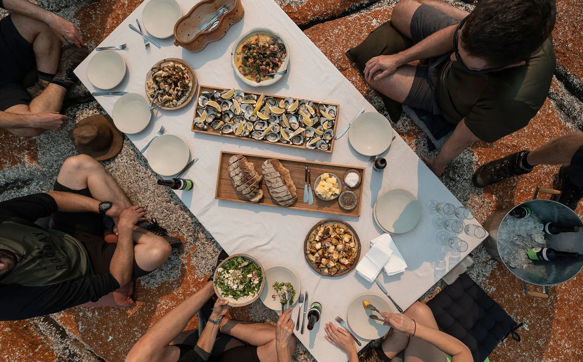 A top-down view of a group around a picnic table perched on an orange-tinged rock. The table is full of oysters, fish and other plates and drinks.