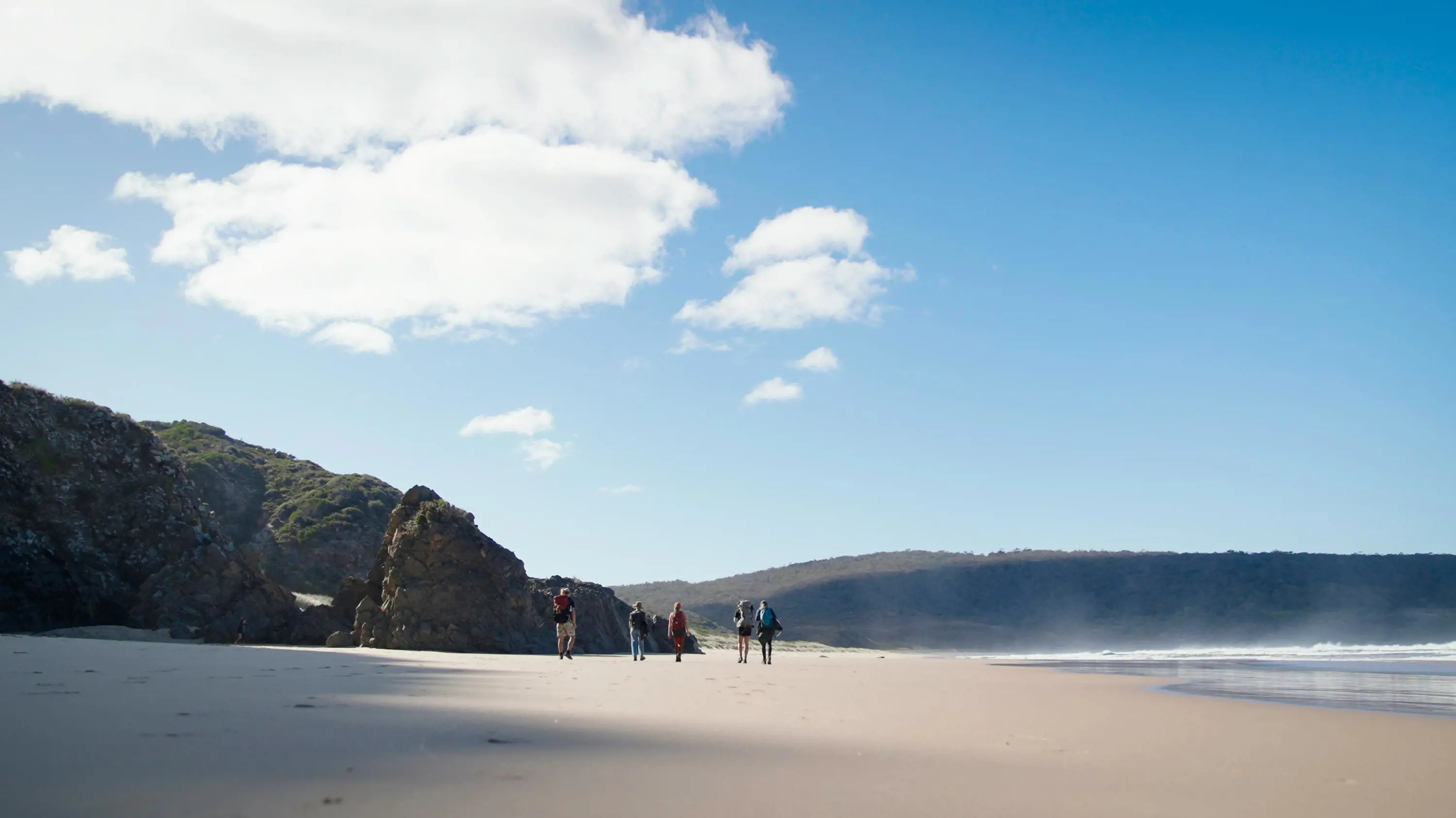 A group of people in the distance walk along a wide beach, with steeply rising bush landscape in the distance.