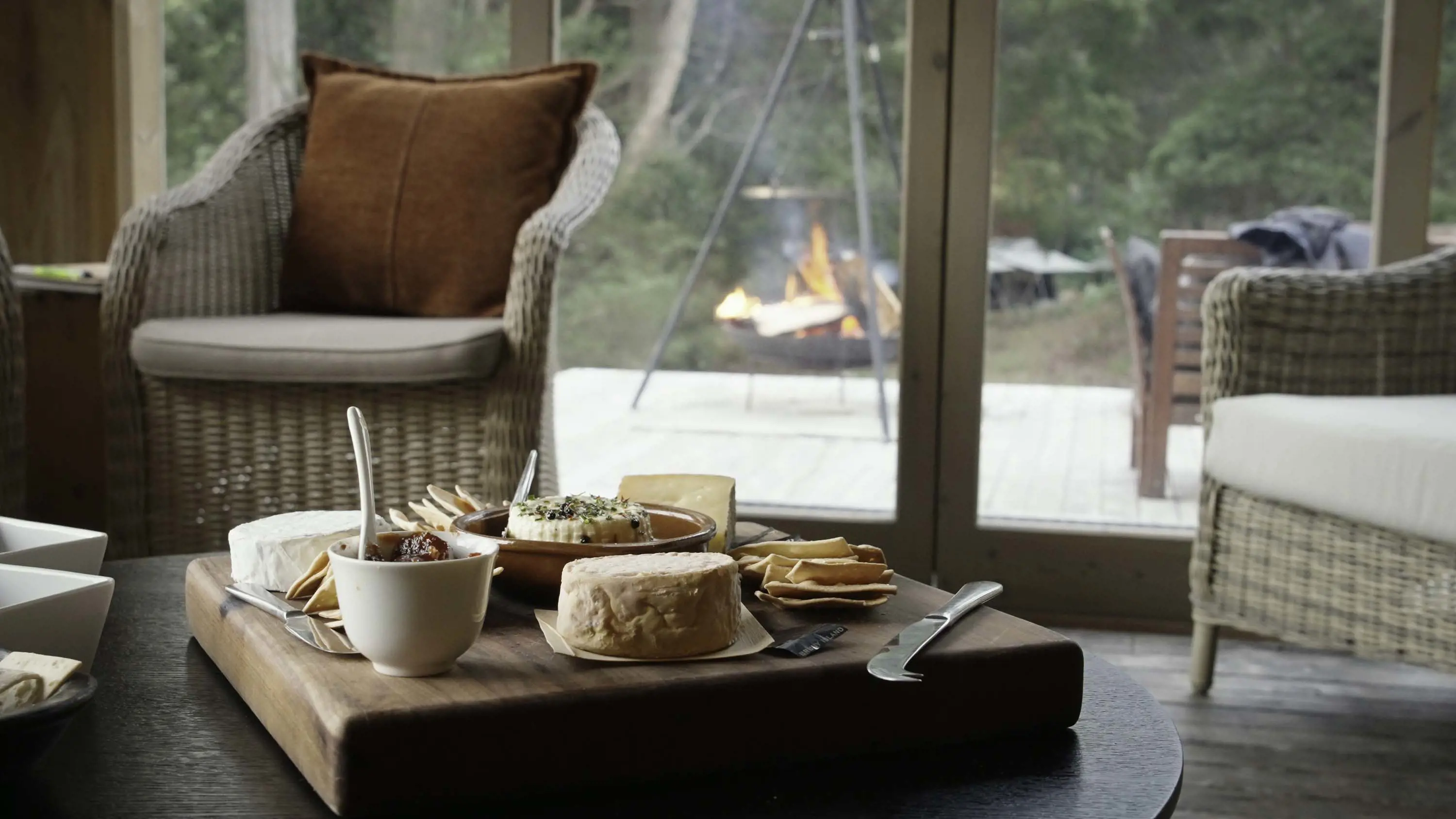 A large wooden charcuterie board on a low table in a loungeroom. Outside, a small hanging firepit is lit.