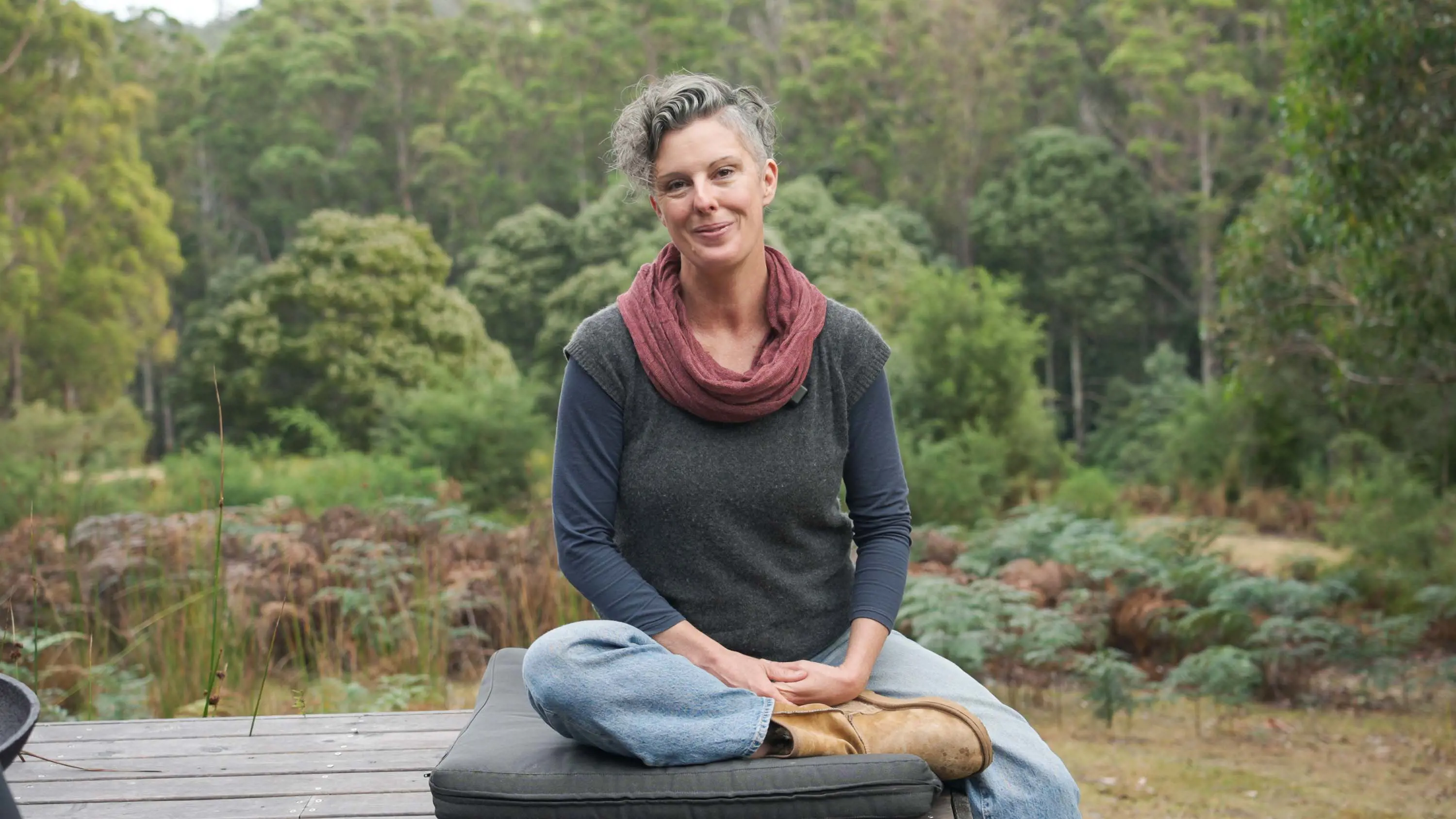 A woman with short hair and a red scarf sits on a cushion on a wooden deck. Behind her is the bush. 