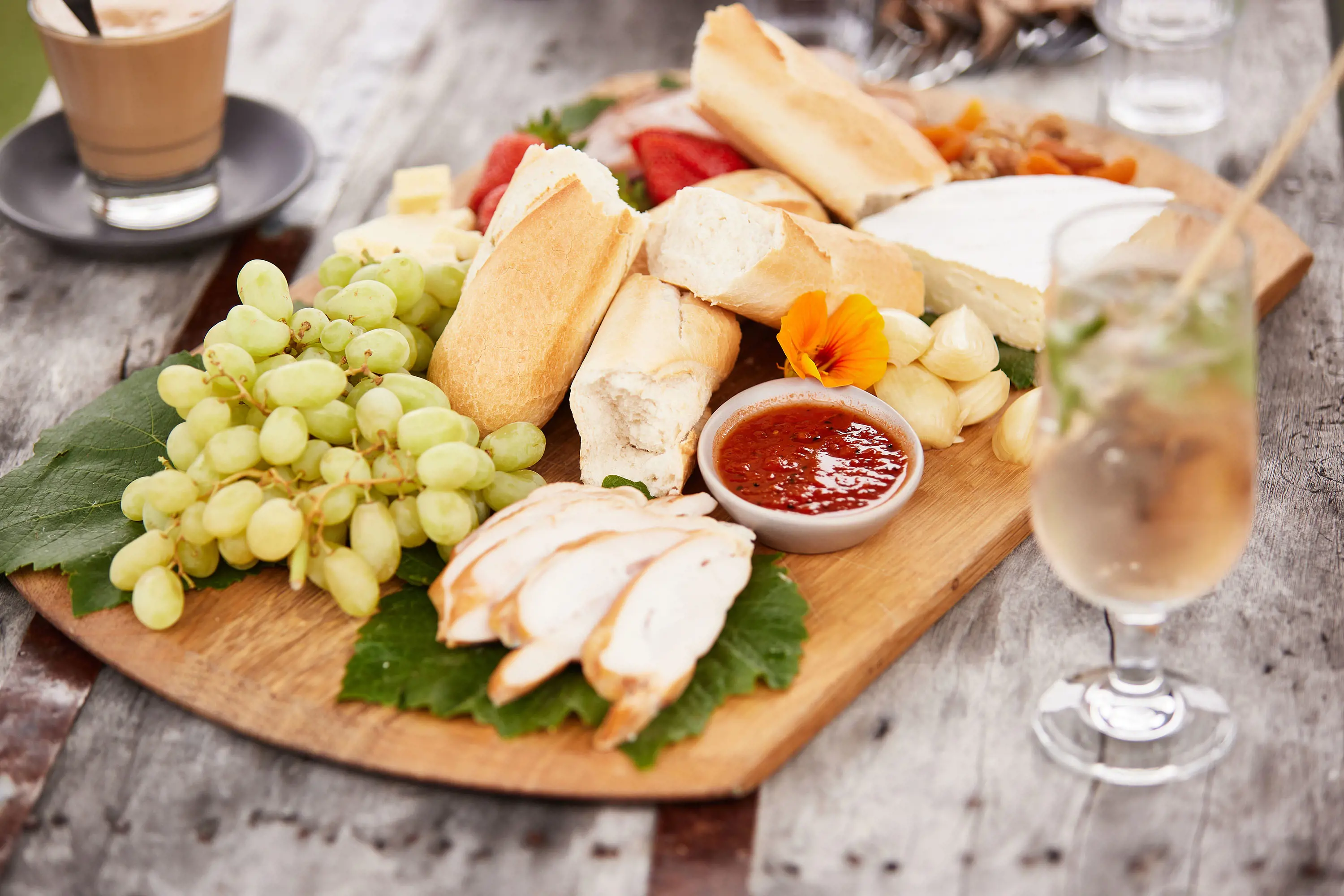 A grazing platter with grapes, bread, cheeses, sliced meats, garlic cloves and strawberries, on an outdoor wooden table next to some drinks.