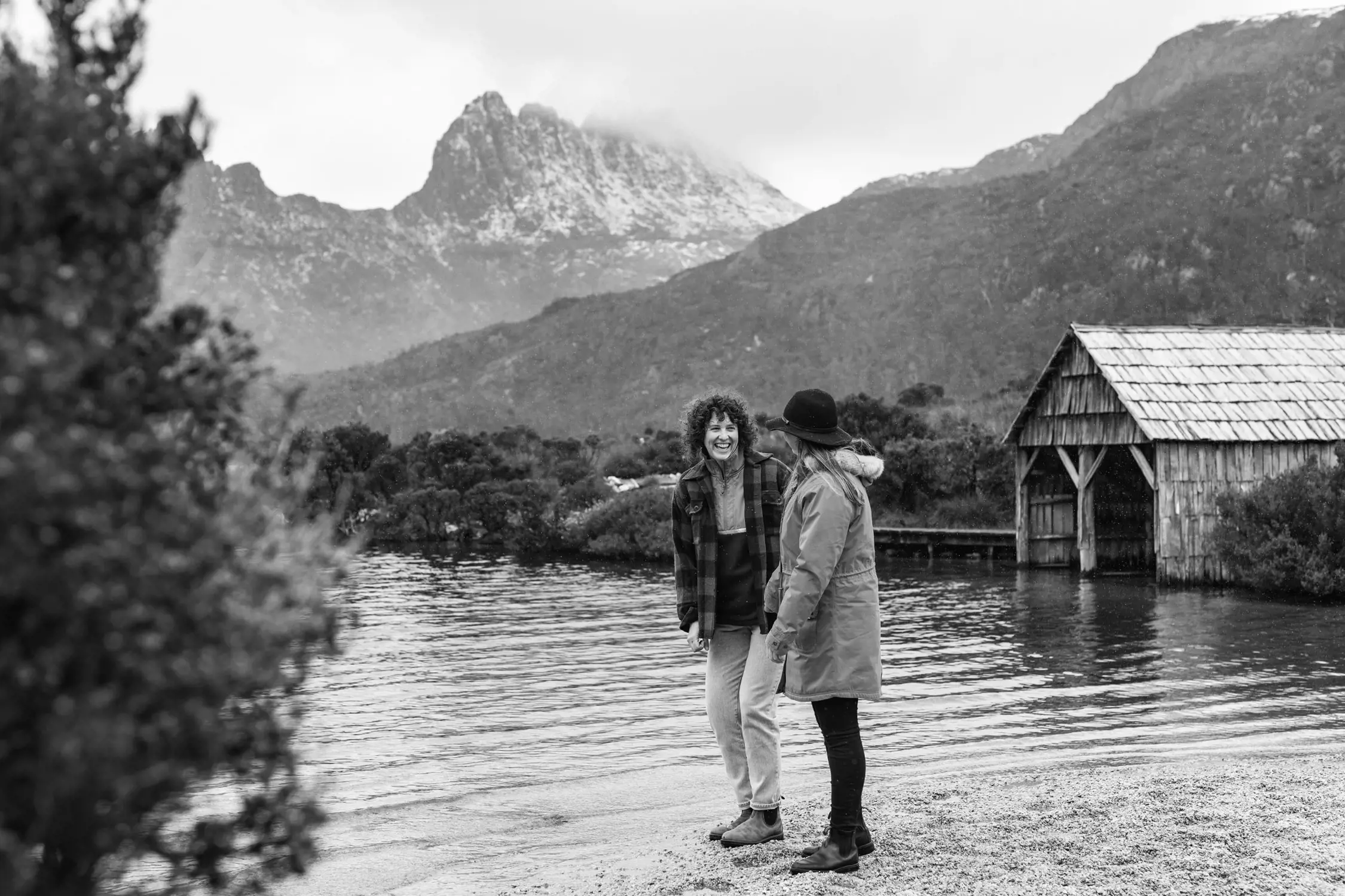 Two women are standing near the edge of a calm lake, laughing together. One is wearing a plaid jacket, and the other is dressed in a warm coat and hat. Behind them, mist-covered mountains rise dramatically into the sky, and a rustic wooden boat shed sits at the water's edge. The scene exudes a sense of joy and connection in a beautiful natural environment