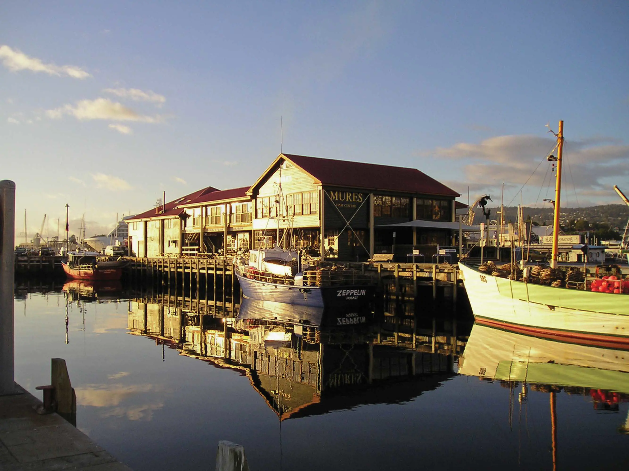 A large two-story building stands on the edge of a concrete wharf, reflected in the still water below.