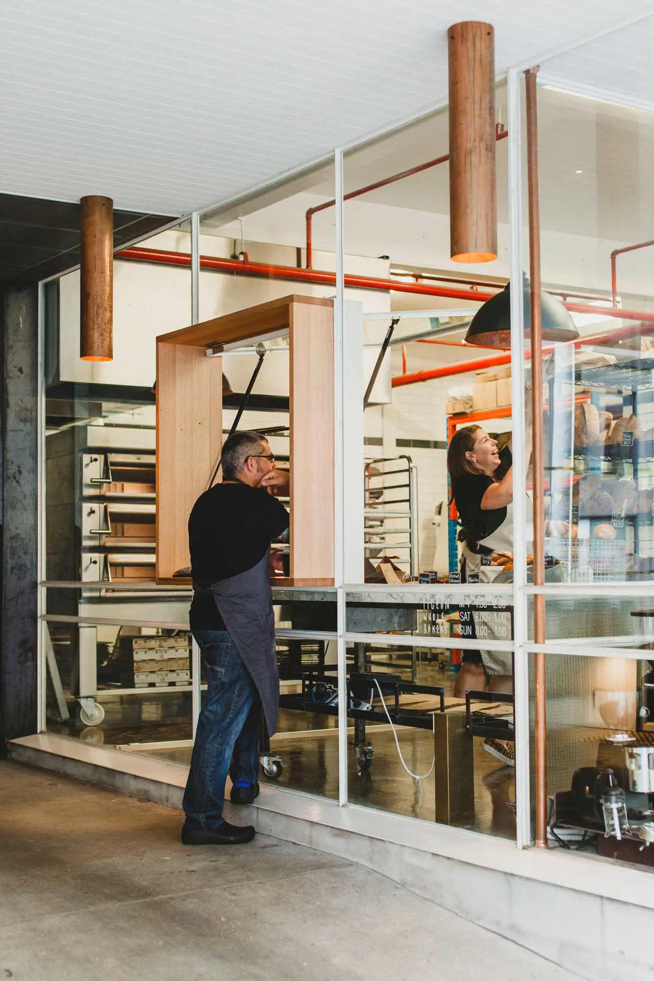 A baker speaks with a colleague through a open window into a bakery space filled with freshly baked bread.