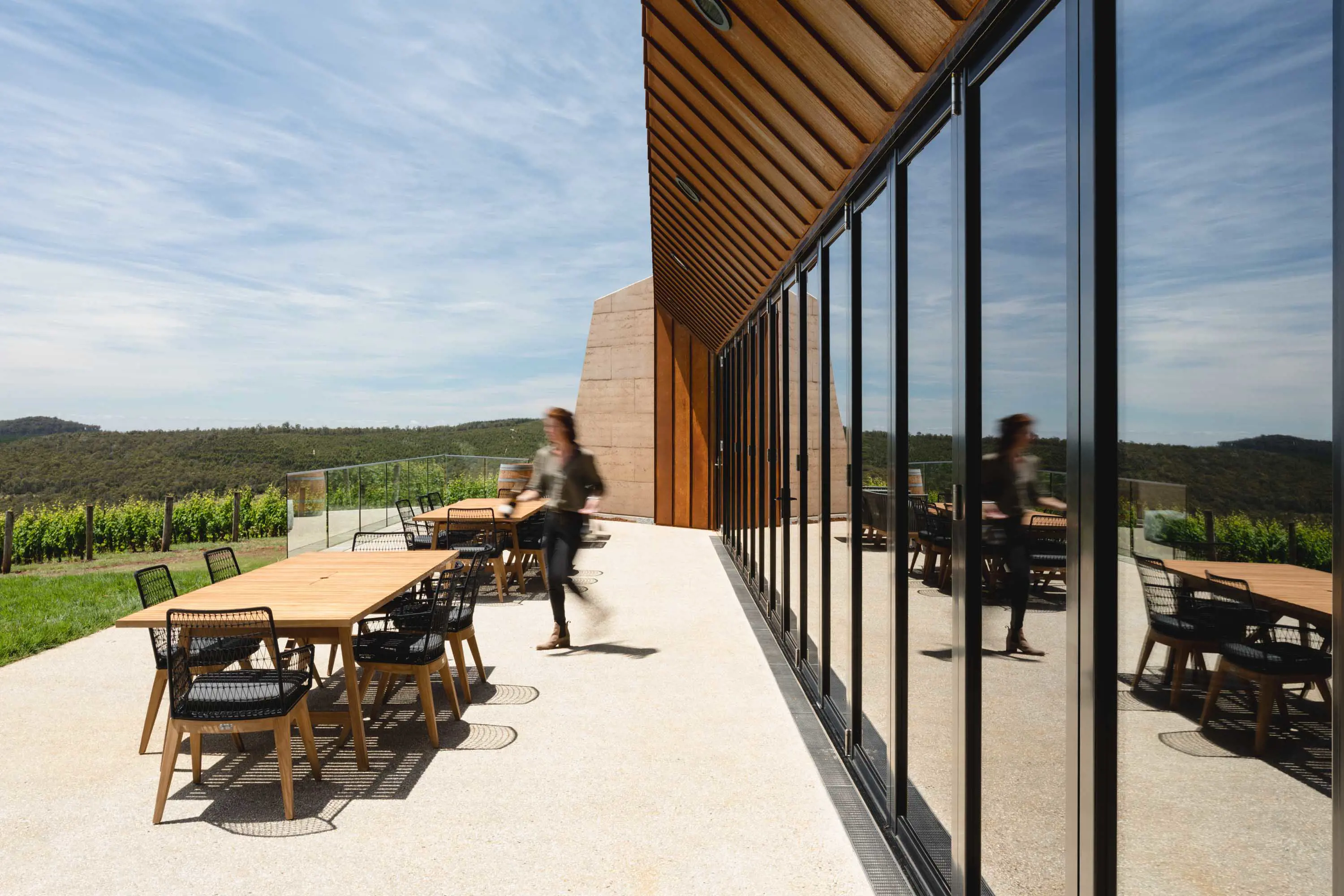 A person walks to a table in the seating area outside a contemporary building with large glass windows looking over a vienyard.