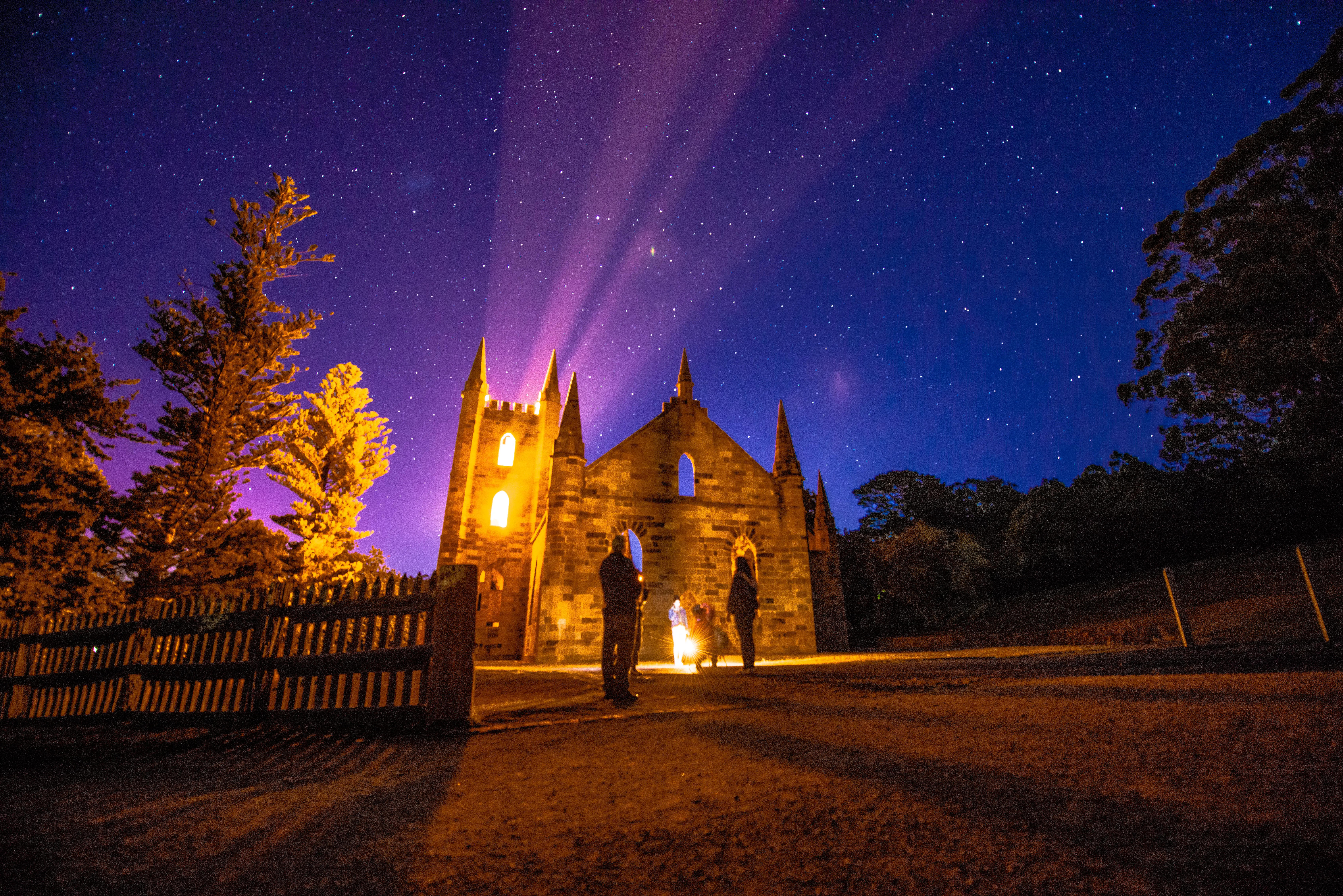 A church, illuminated from within with warm light contrasts with the purple sky filled with stars. A group of people stand in front of the entrance and look up.