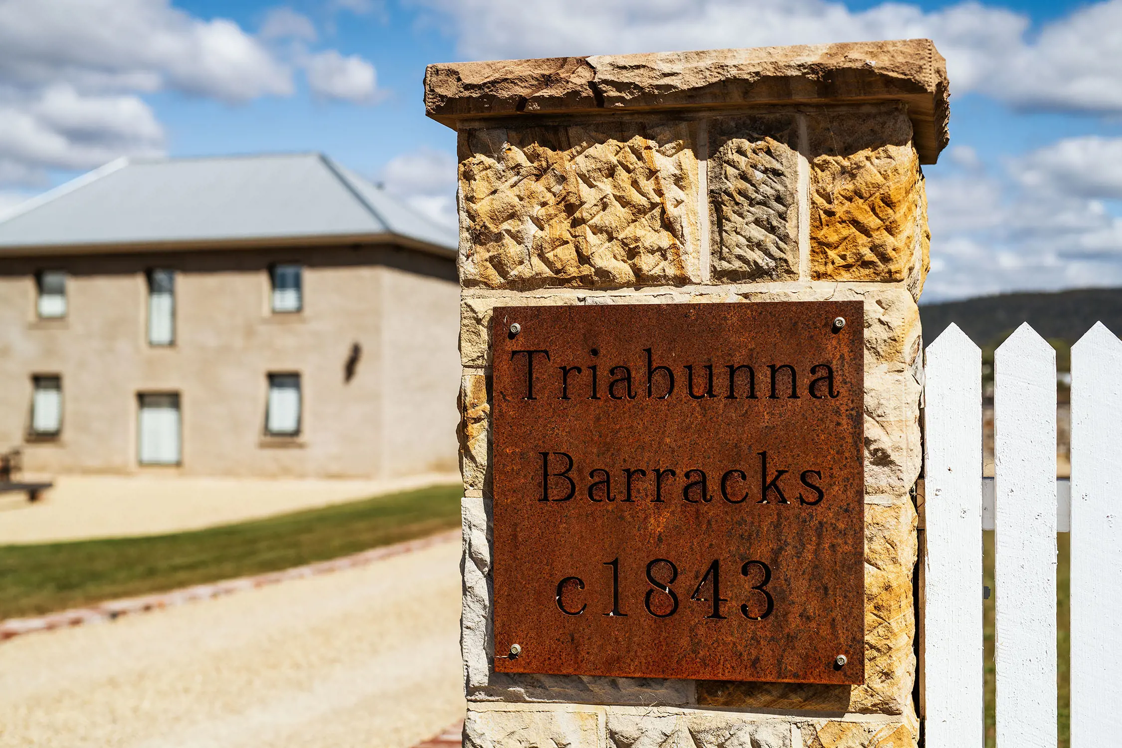 A stylistically rusted metal plate on a sandstone pillar in front of a neatly manicured heritage building facade. The sign reads "Triabunna Barracks c 1843".