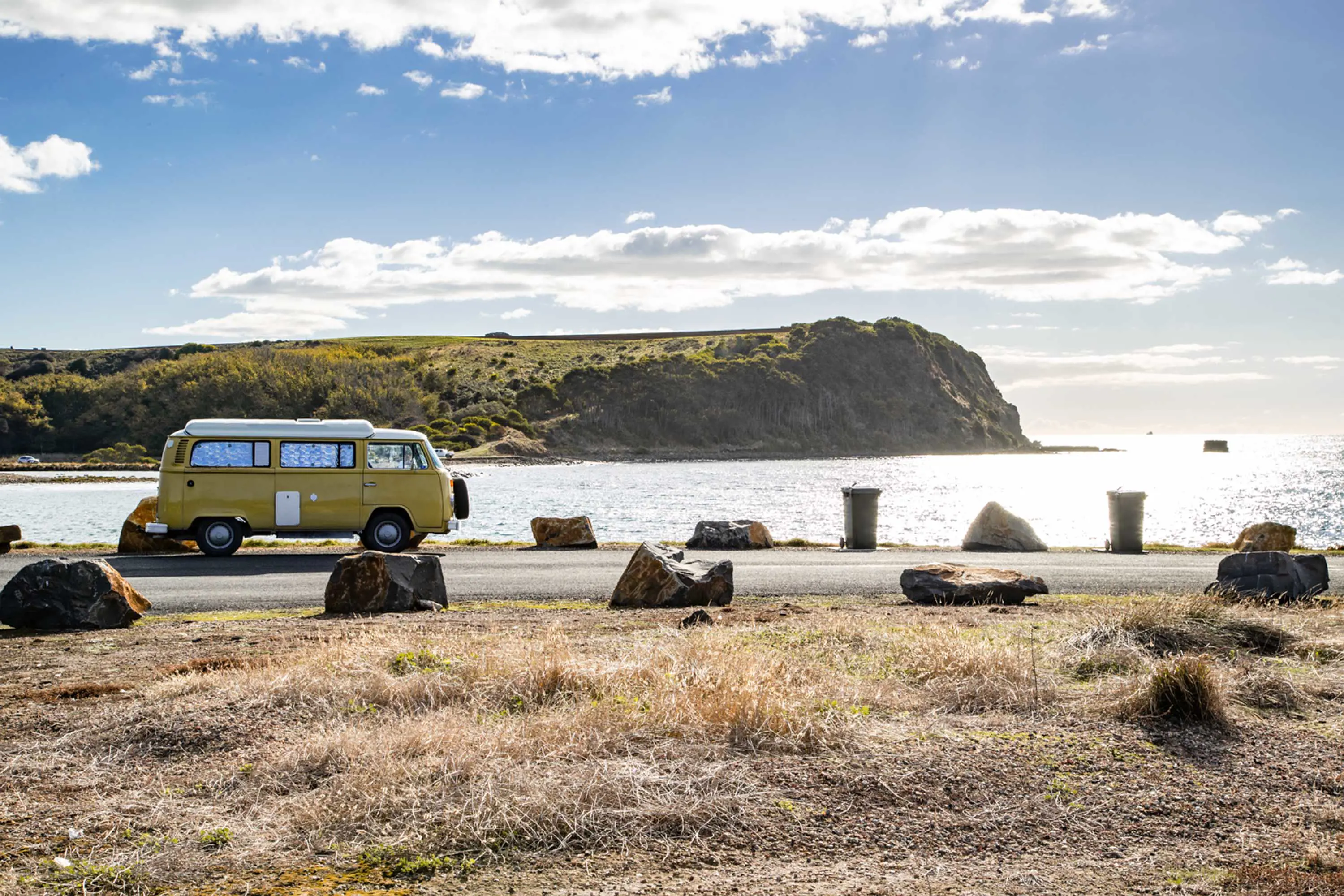 A yellow vintage van with a white roof parks on a road by the side of an inlet. A rocky peninsula in the distant background.