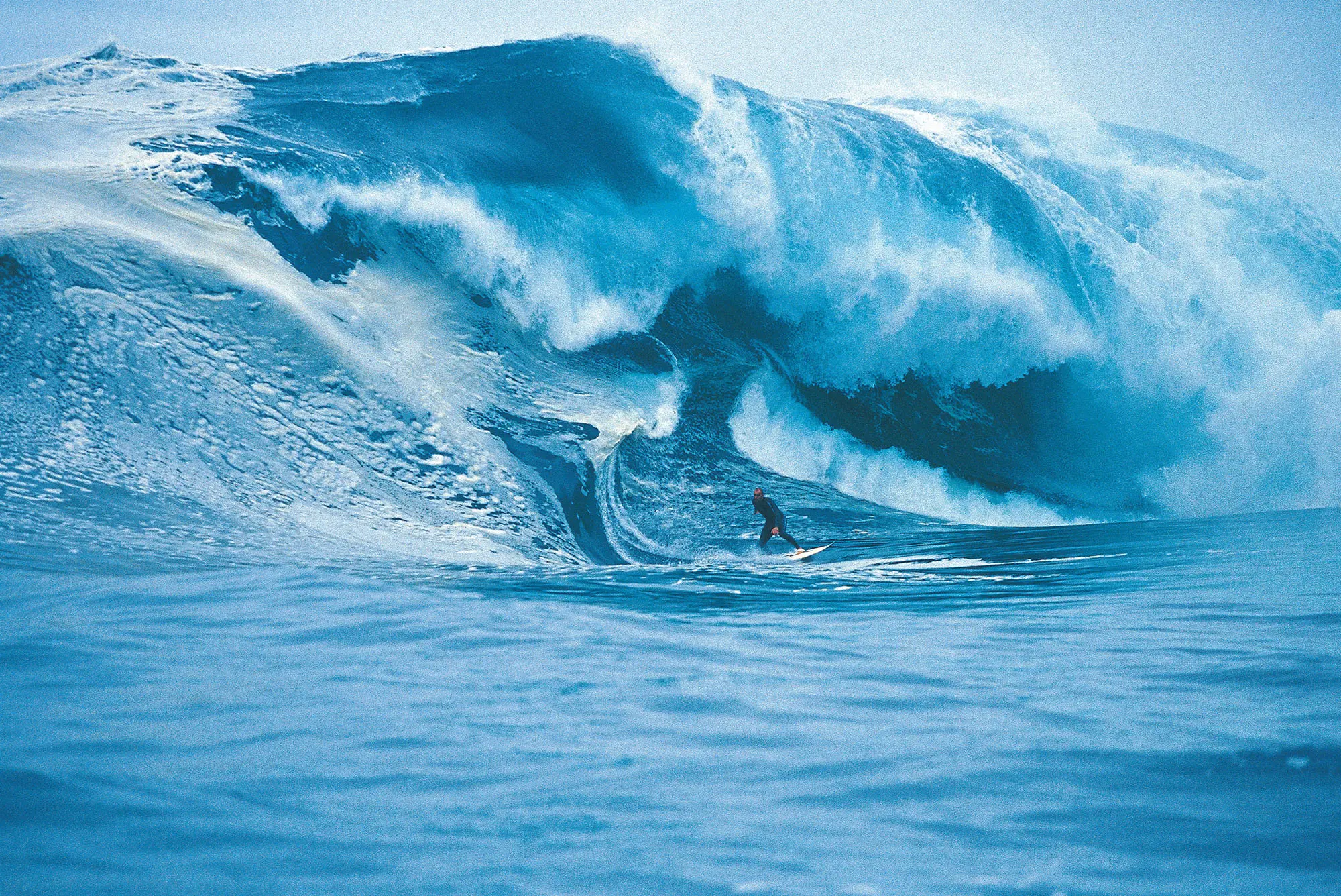 A wetsuit-clad person on a surfboard is dwarfed by a huge, undulating, richly blue wave he is surfing.