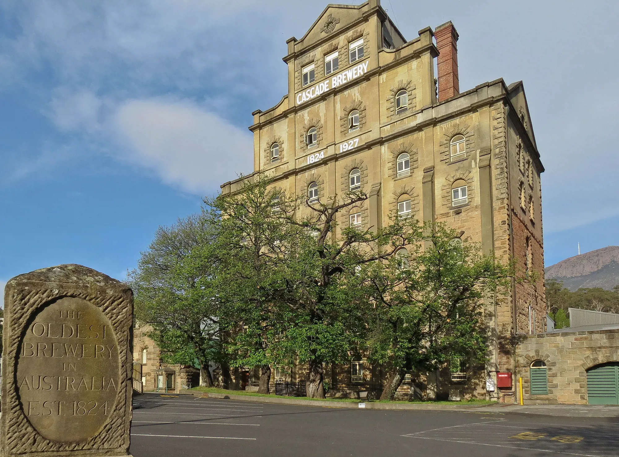 A sandstone plinth saying "The oldest brewery in Australia - est. 1824" sits in front of a large multistorey sandstone building rising from some trees. The white sign atop the building says "Cascade Brewery".