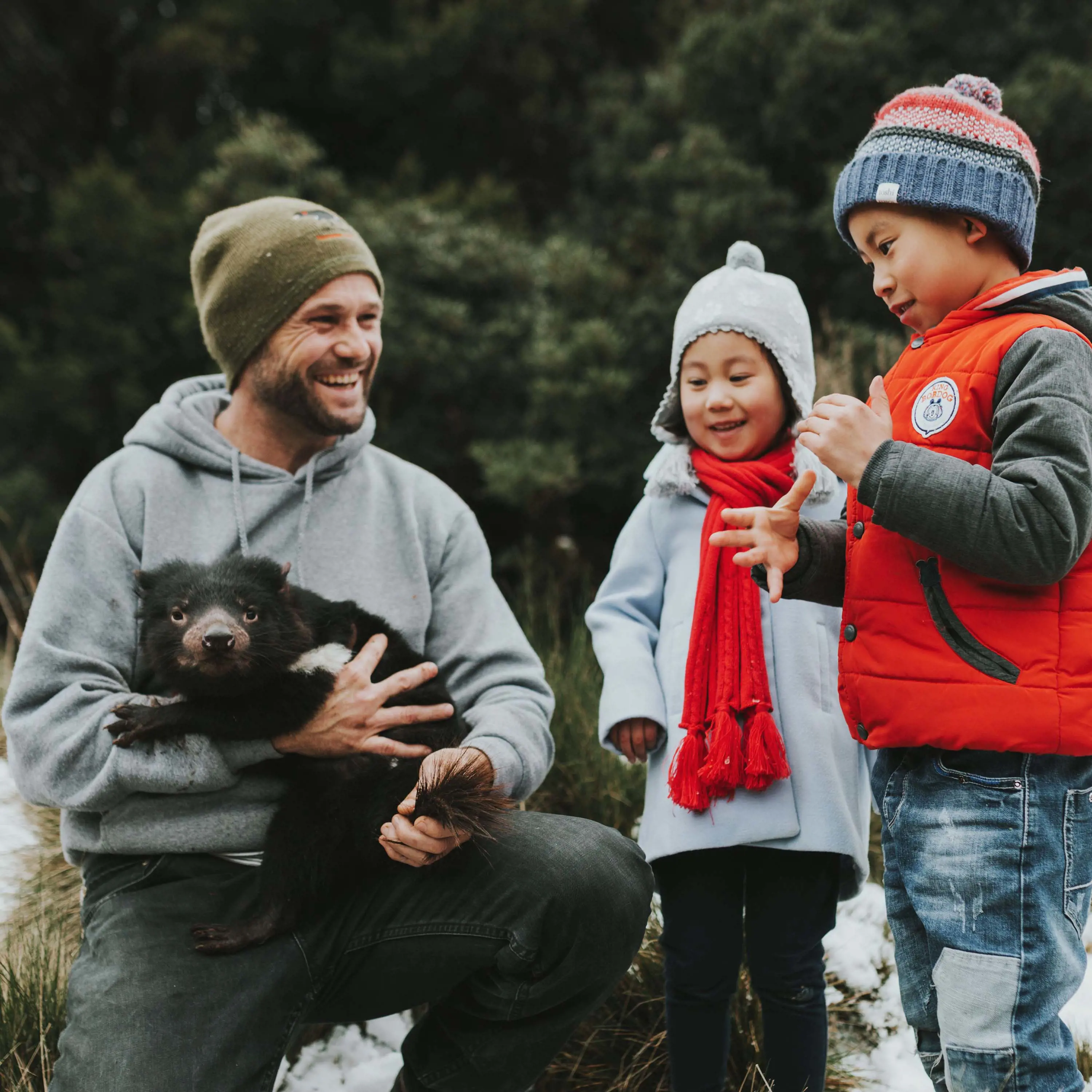 A man kneeling in the snow is holding a Tasmanian devil in his arms and grinning to two children, who are entranced by the creature.