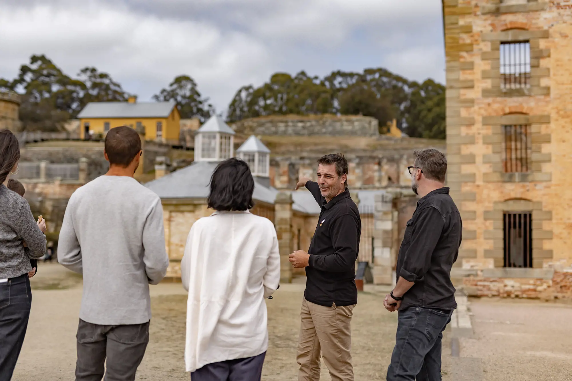 A tour group stand listening to a guide, who is speaking and pointing towards a row of sandstone heritage buildings.