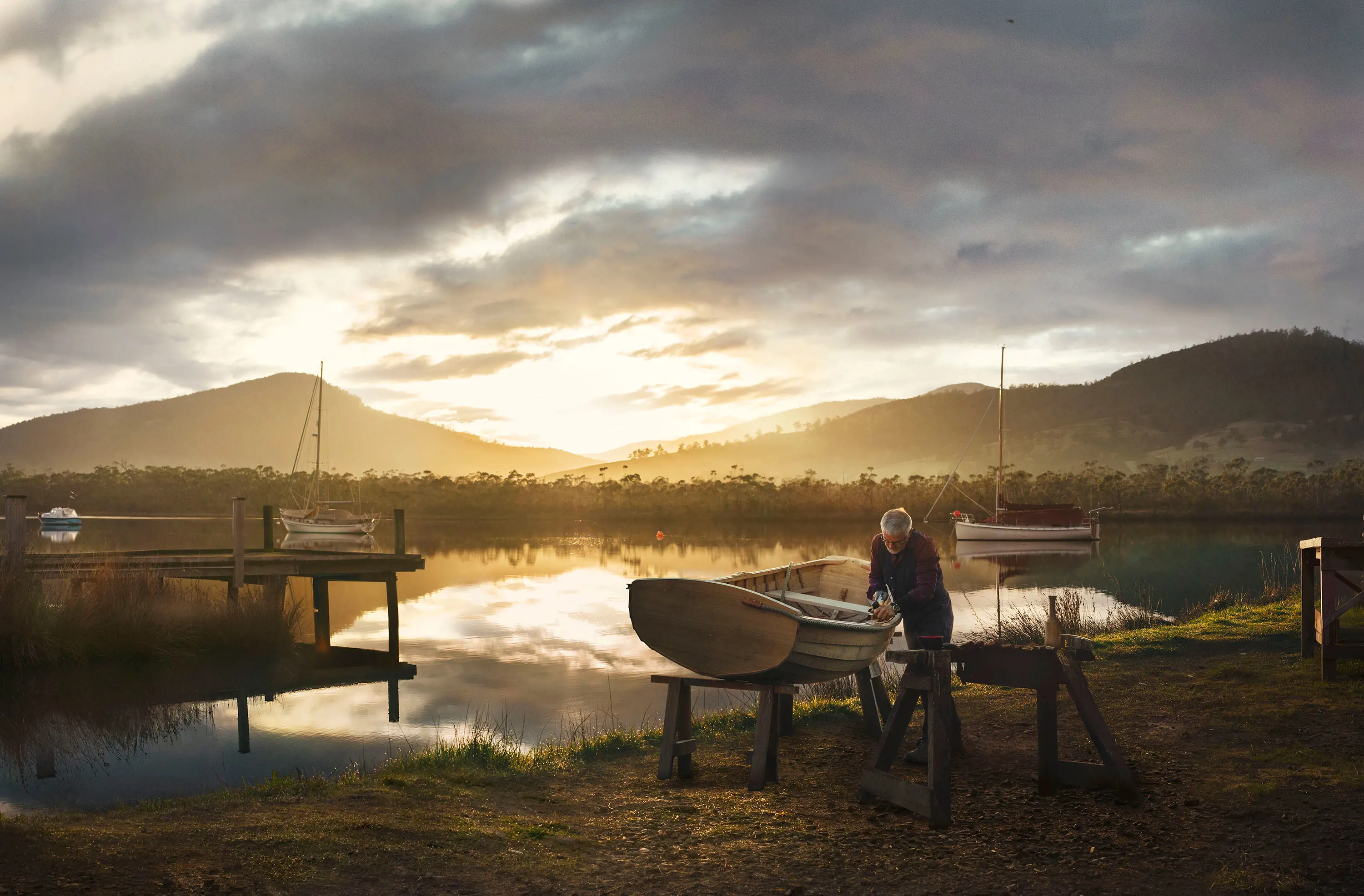 By the banks of a still, picturesque river, a man works on a wooden dinghy propped up on two sawhorses.