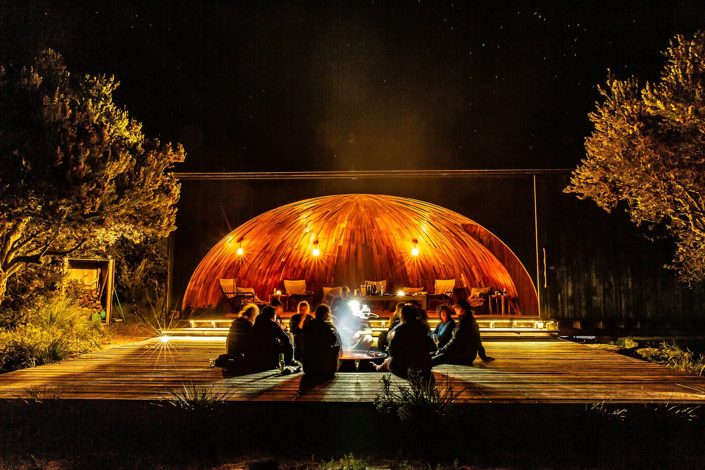 On a raised wooden deck, in front of a curved half-dome, a group of people sit around a fire pit. The night sky is inky black and the lights are illuminating the trees beside the platform.