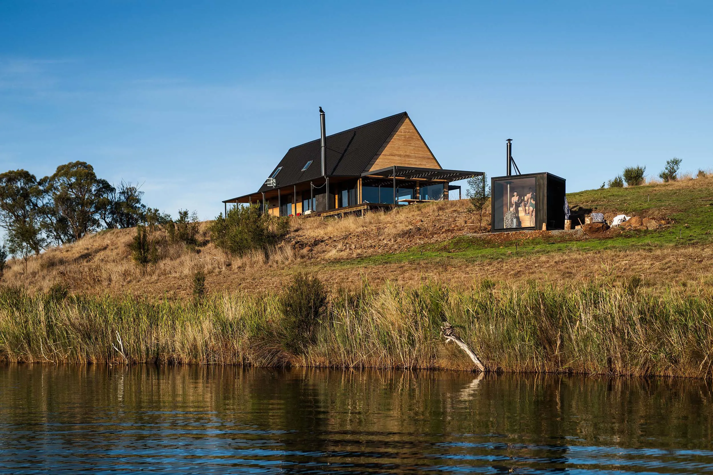 A wooden house with a sleek black roof and walls of windows overlooks a small black sauna set into the hillside above a river.