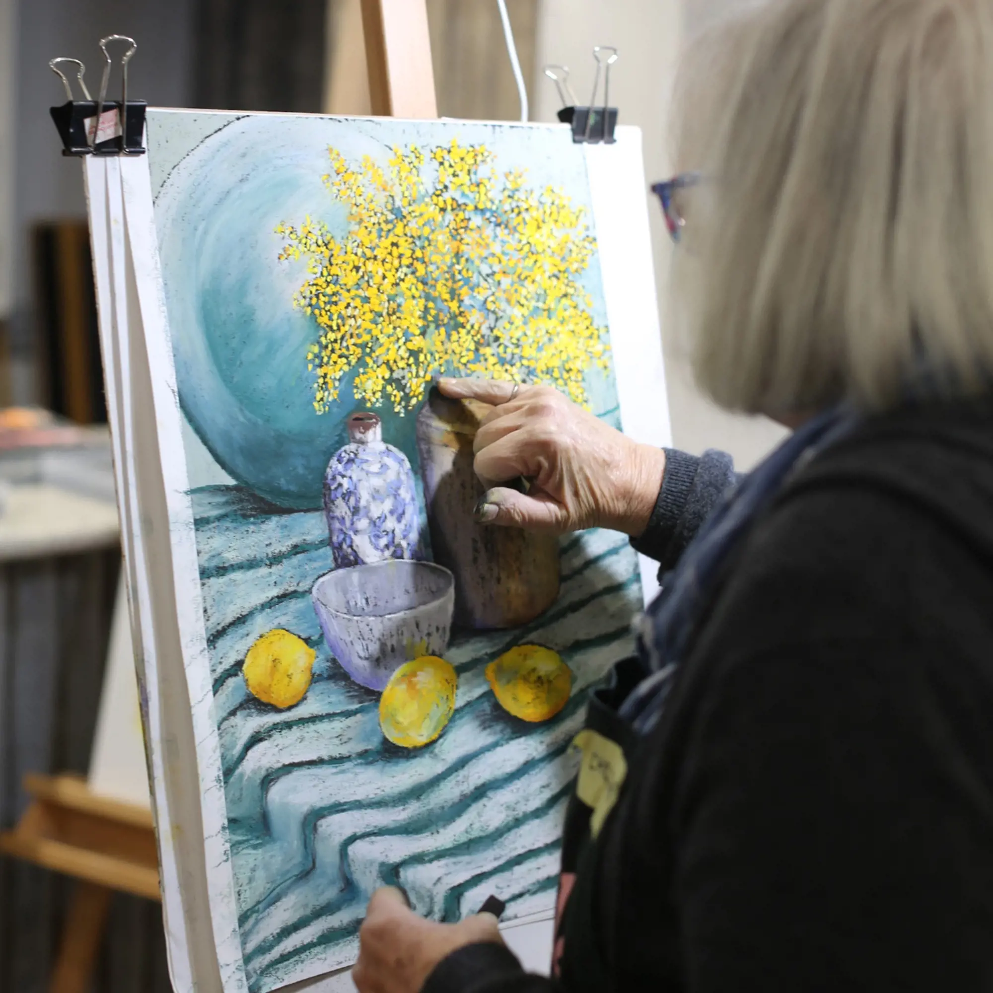 A woman uses her pinky finger to blend some colours on a work in progress of a still life.