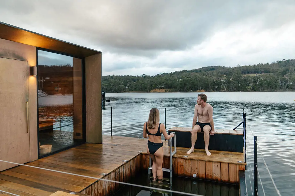 A sleek wooden sauna boat floats on a river. Two people are on the deck in their swimmers, climbing out of the water.