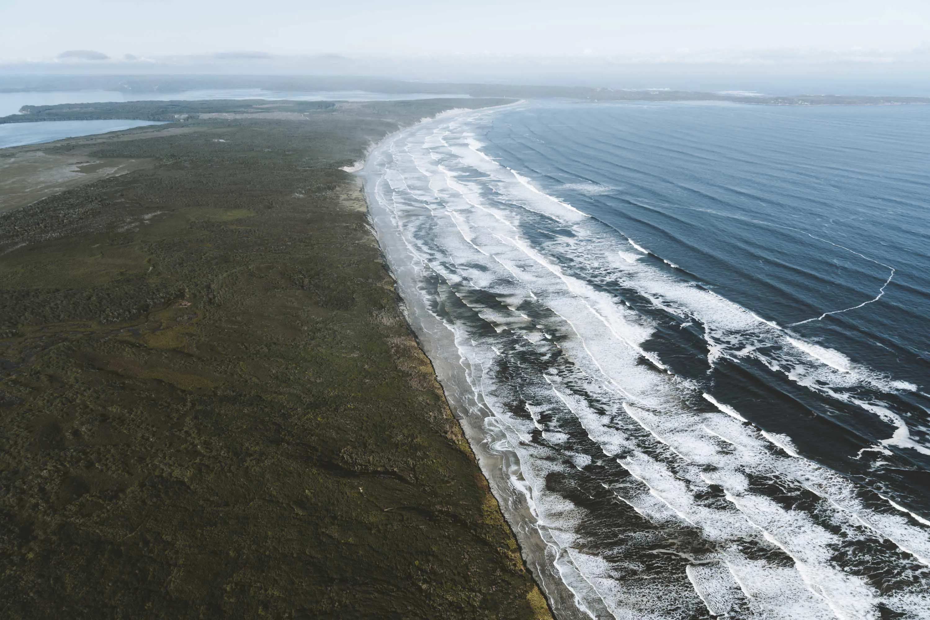 An aerial photograph of a beach with rough waves crashing on a narrow strip of grey sand.
