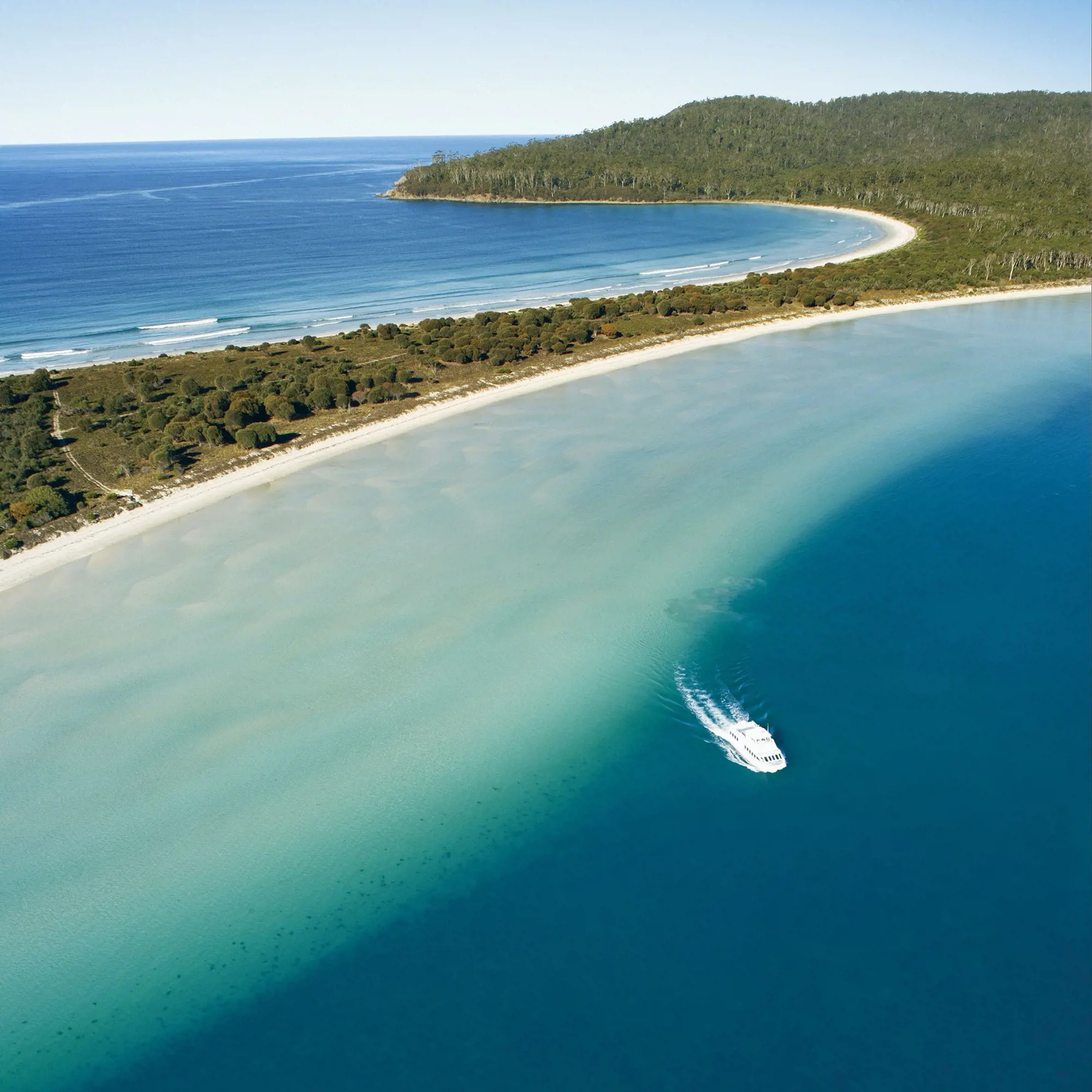 An aerial view of a densely forested island. A narrow isthmus is surrounded by deep blue waters, and a small boat is in one of the bays.