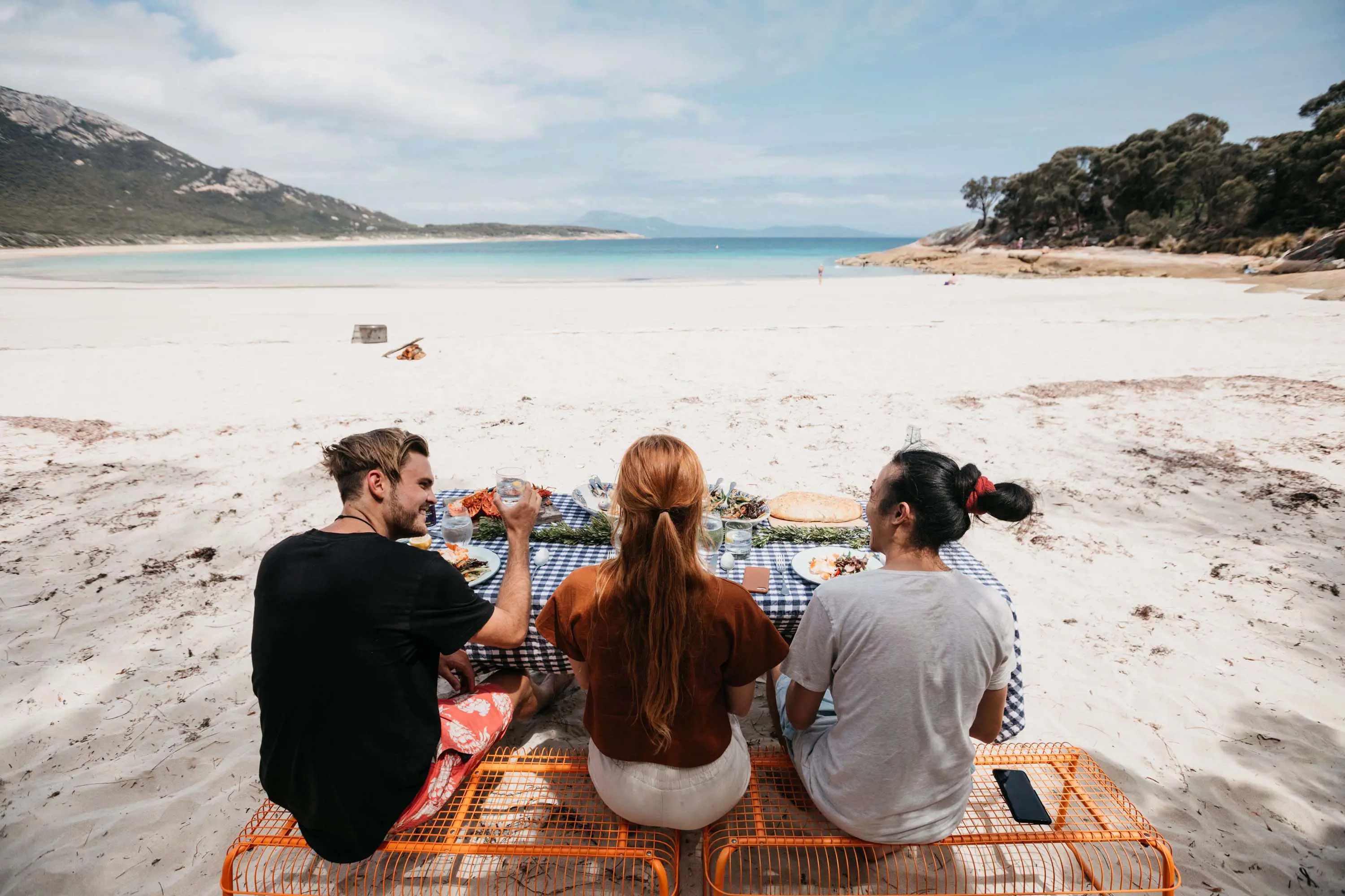 Three people are sitting on the sand at a beautiful sheltered beach, with a picnic blanket spread out filled with food and drinks.