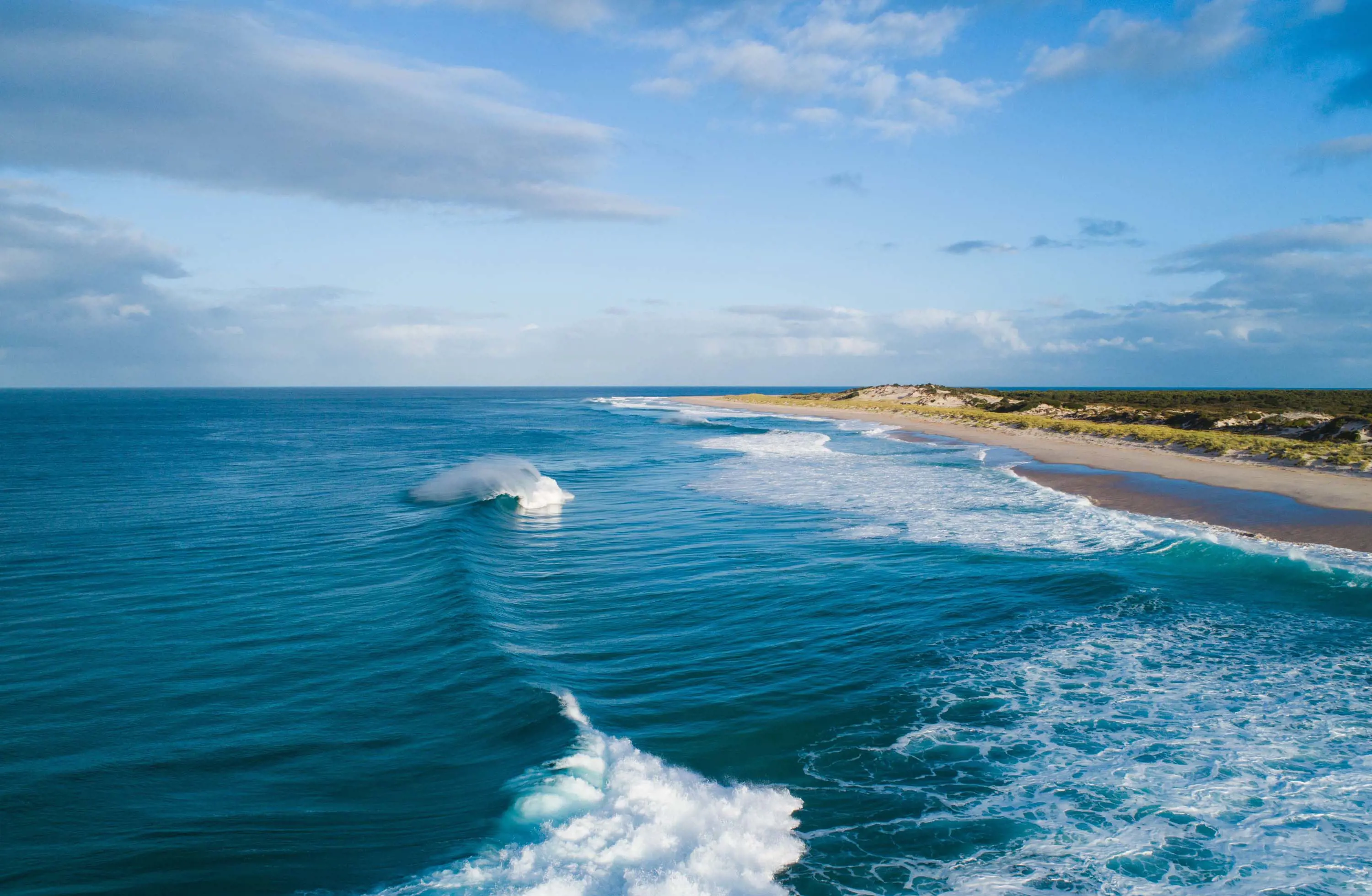 An aerial image of a picturesque beach, with some rolling crashing waves and a small expanse of land behind the sandy beach.