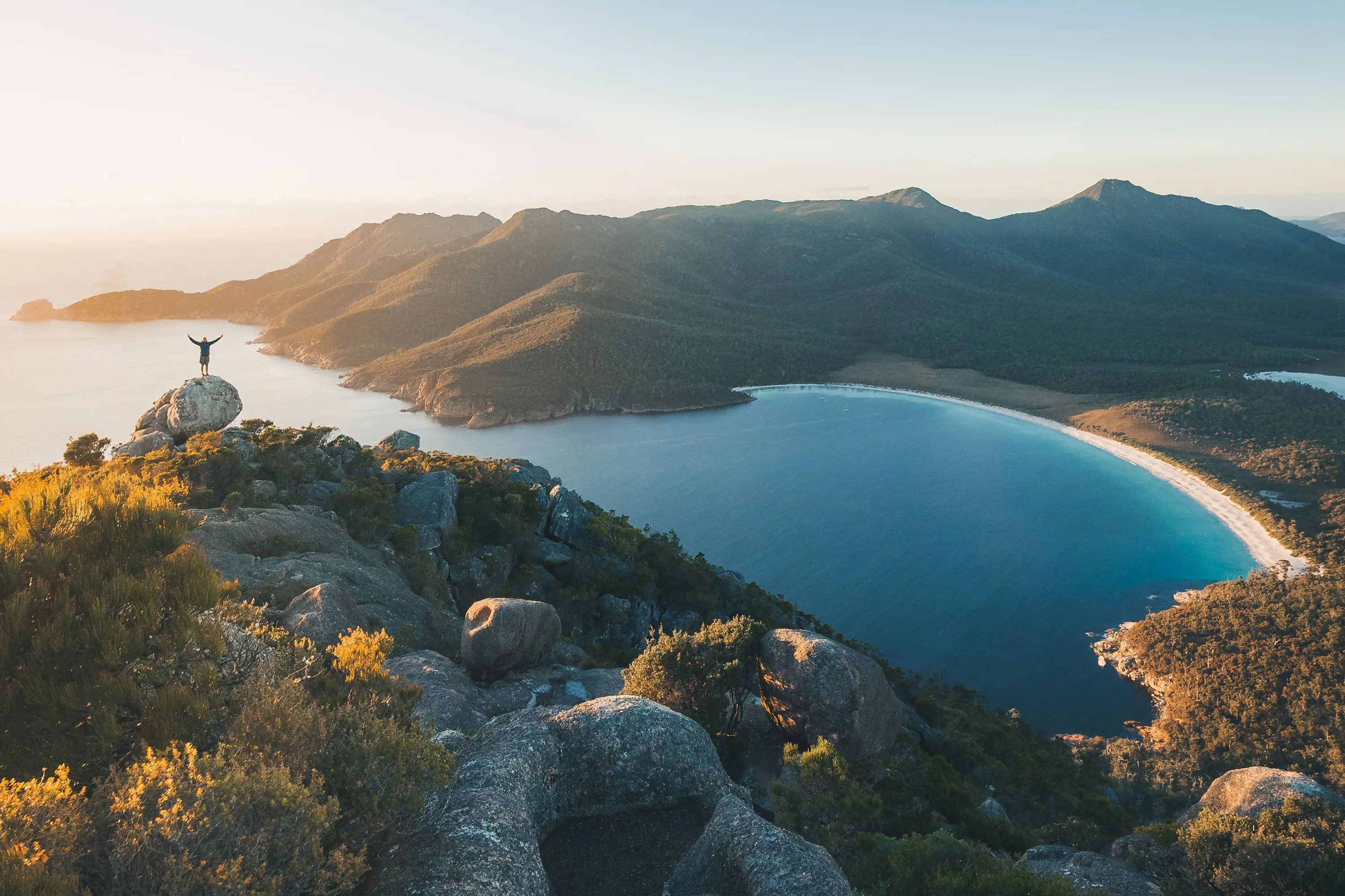 A person stands atop a boulder on a huge cliff, dropping down below to reveal a beautiful ocean bay with a mountainous background.