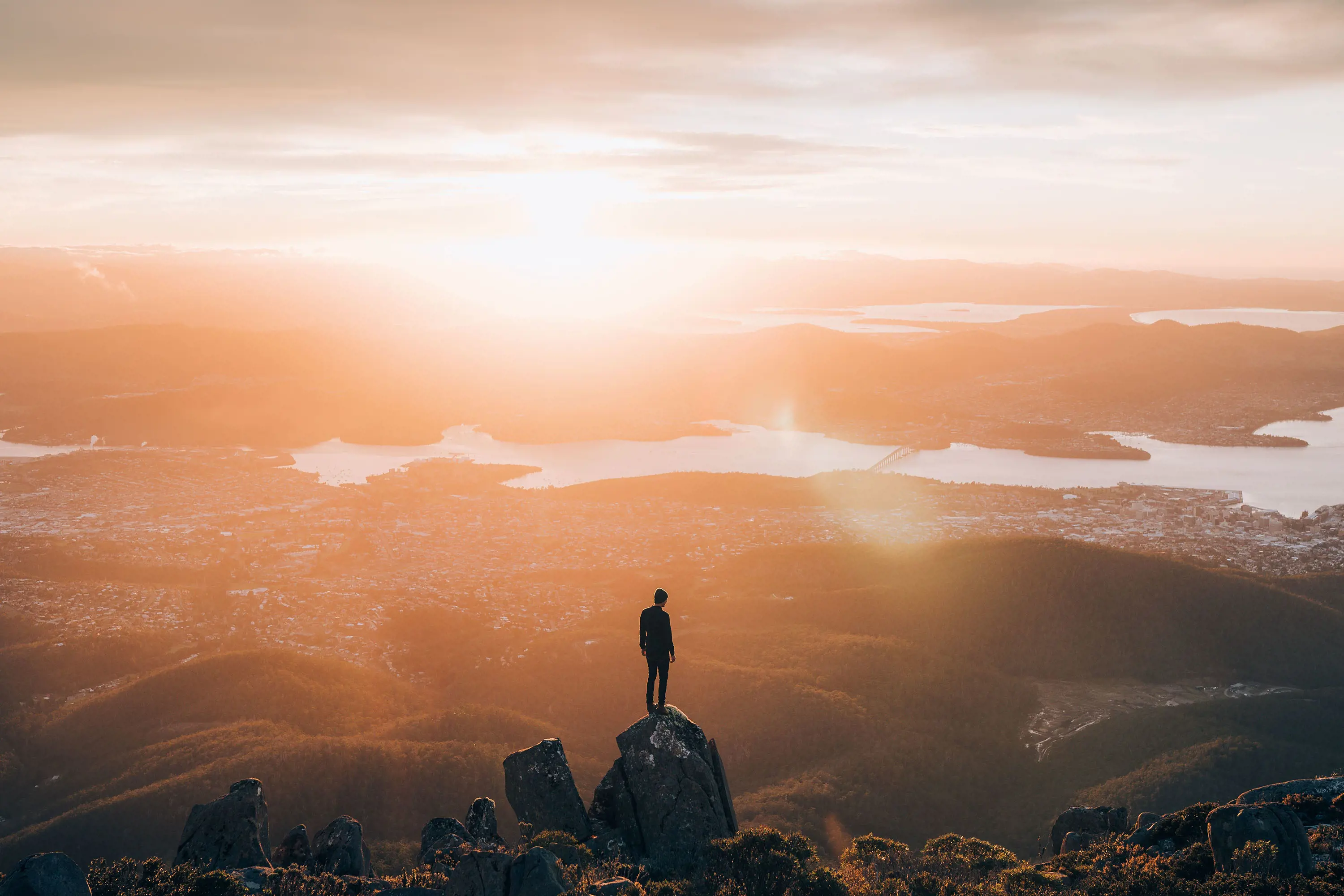 At the top of a mountain, a person stands atop a tall rock, silhouetted by the sun's blare. Below, the city of Hobart sprawls out, following the meandering Derwent River.