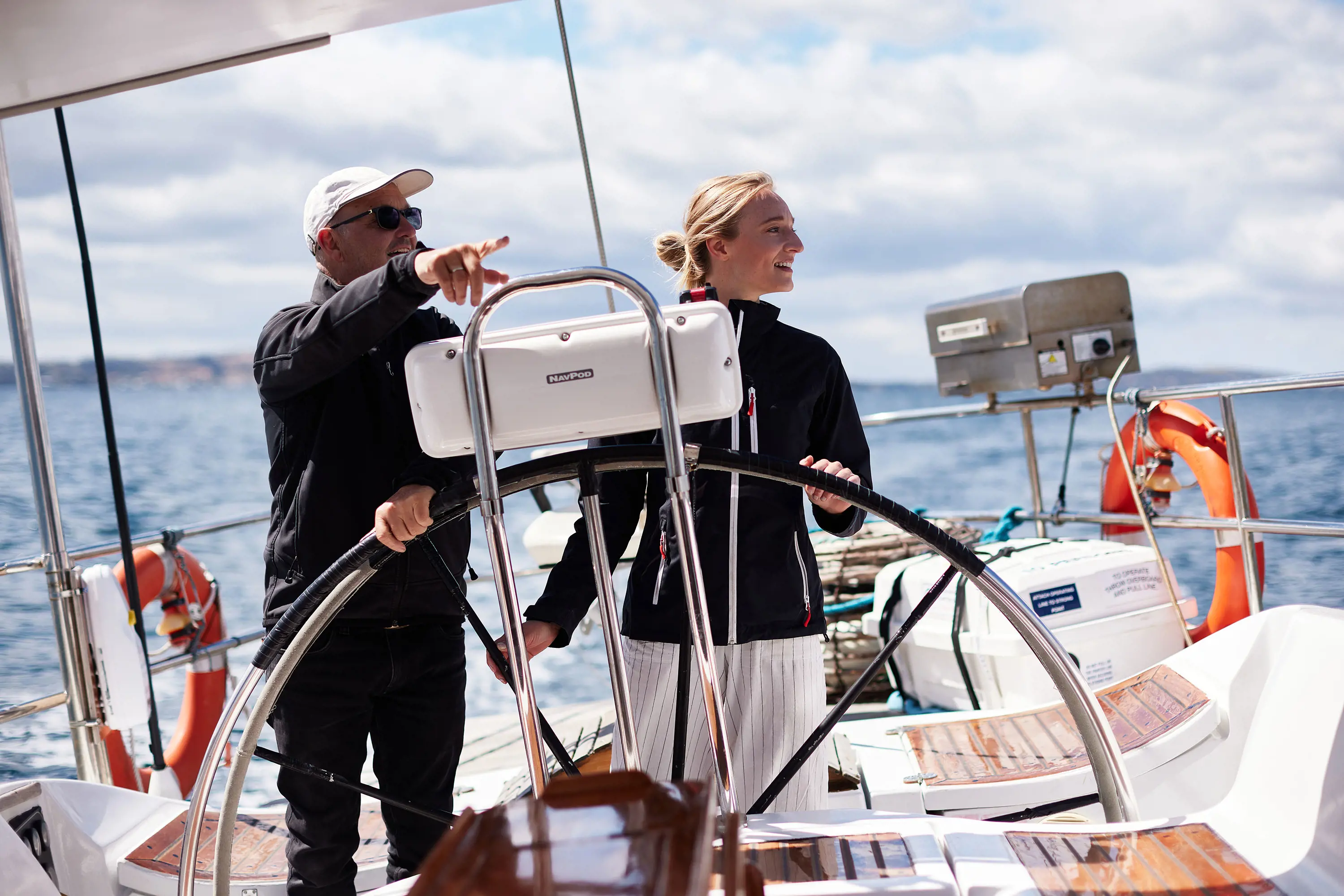 A man points to the water as a woman turns the large metal wheel of a yacht.