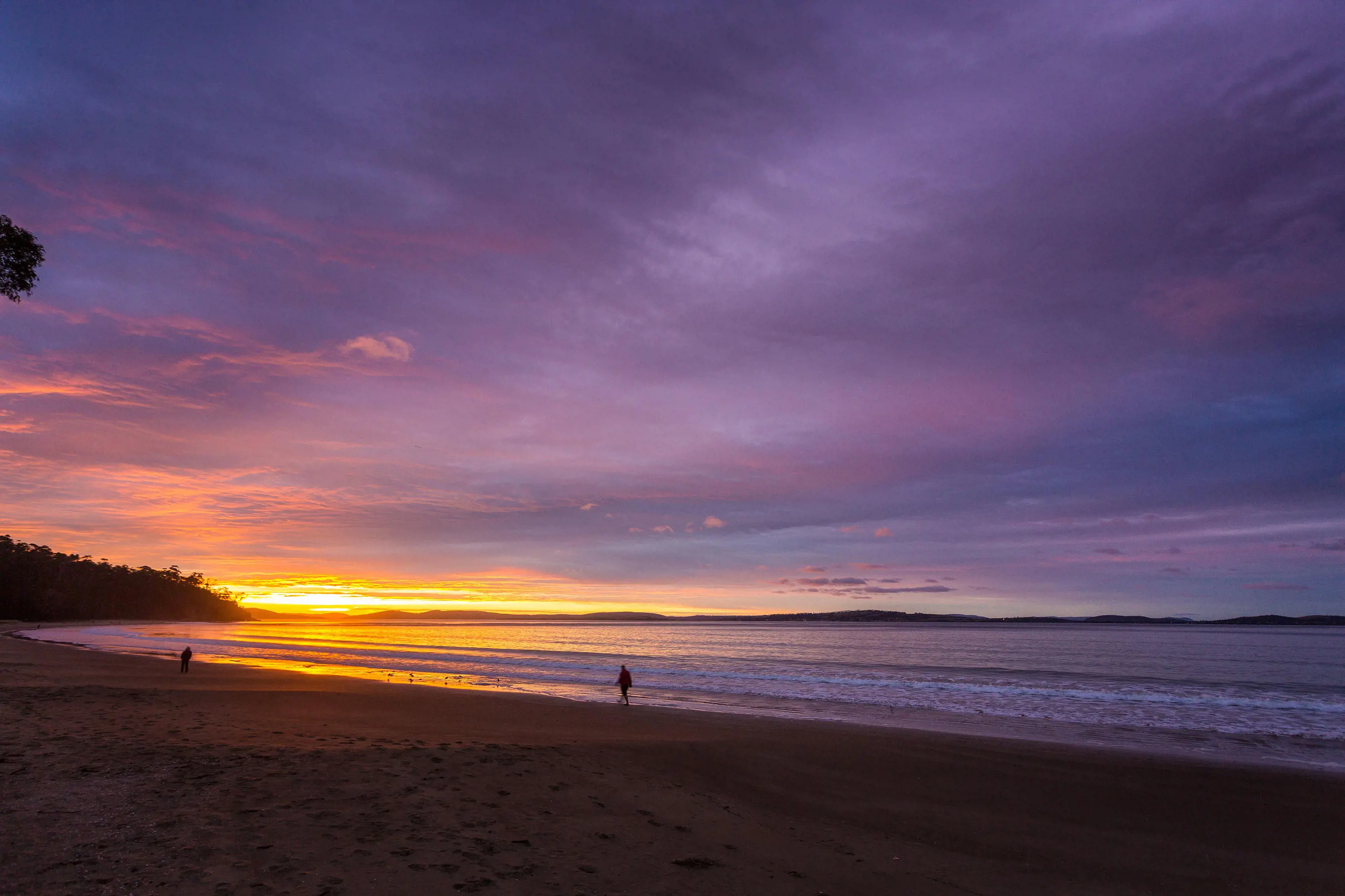 Silhouettes of people walking along a beach. The sky ranges from deep purple, to orange, to bright yellow as the sun rises.