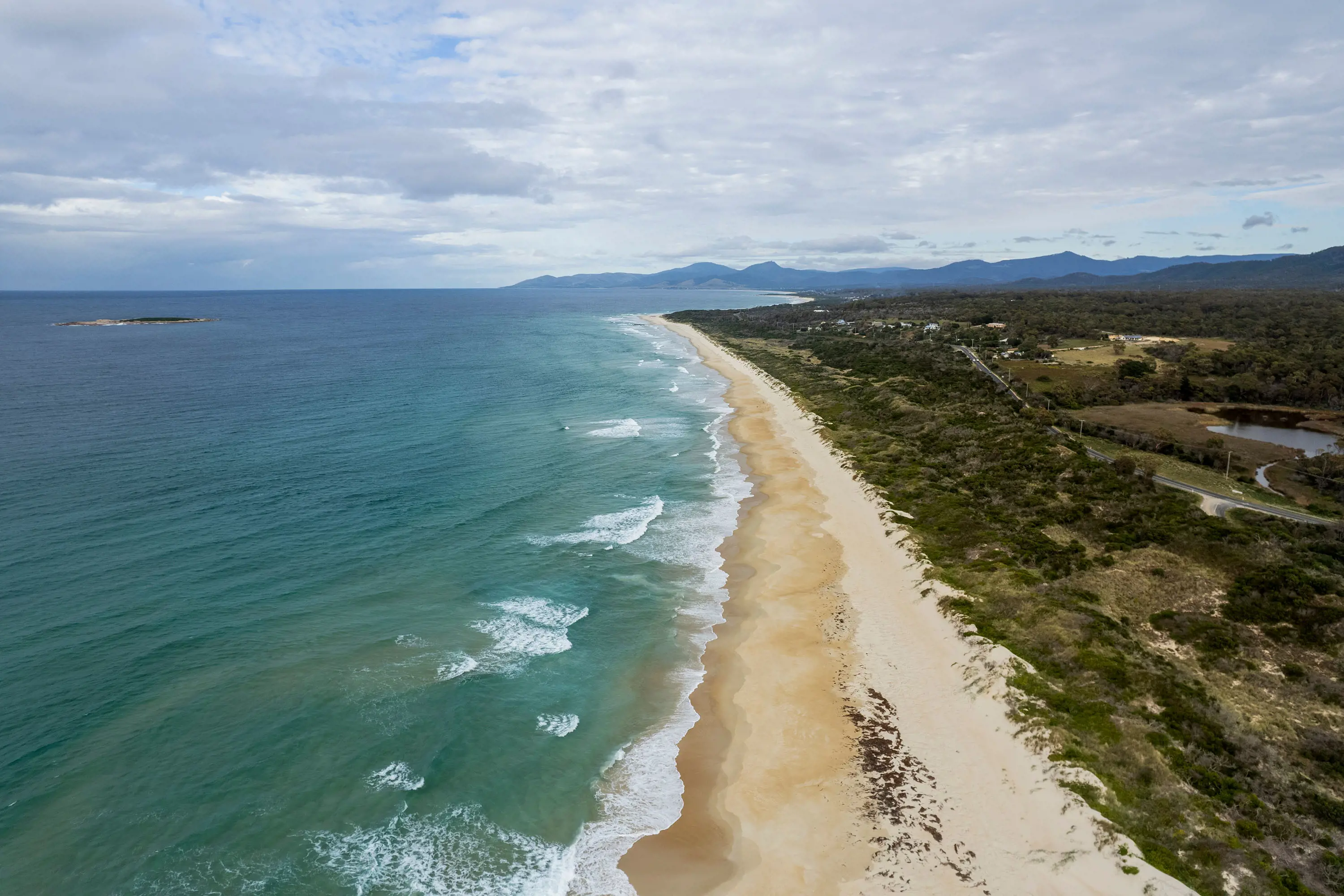 An aerial view, looking down a stretch of beach. Waves roll in on one side of the sand and bush covers the landscape on the other.