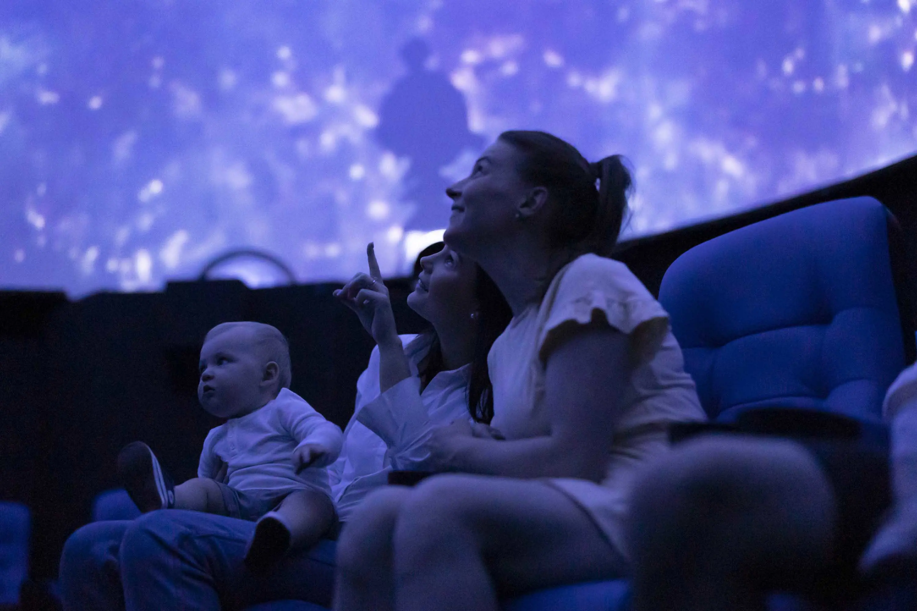 A mother, her daughter and her baby sit in the blue light of a spherical room looking up and pointing at a galaxy projected onto the ceiling.
