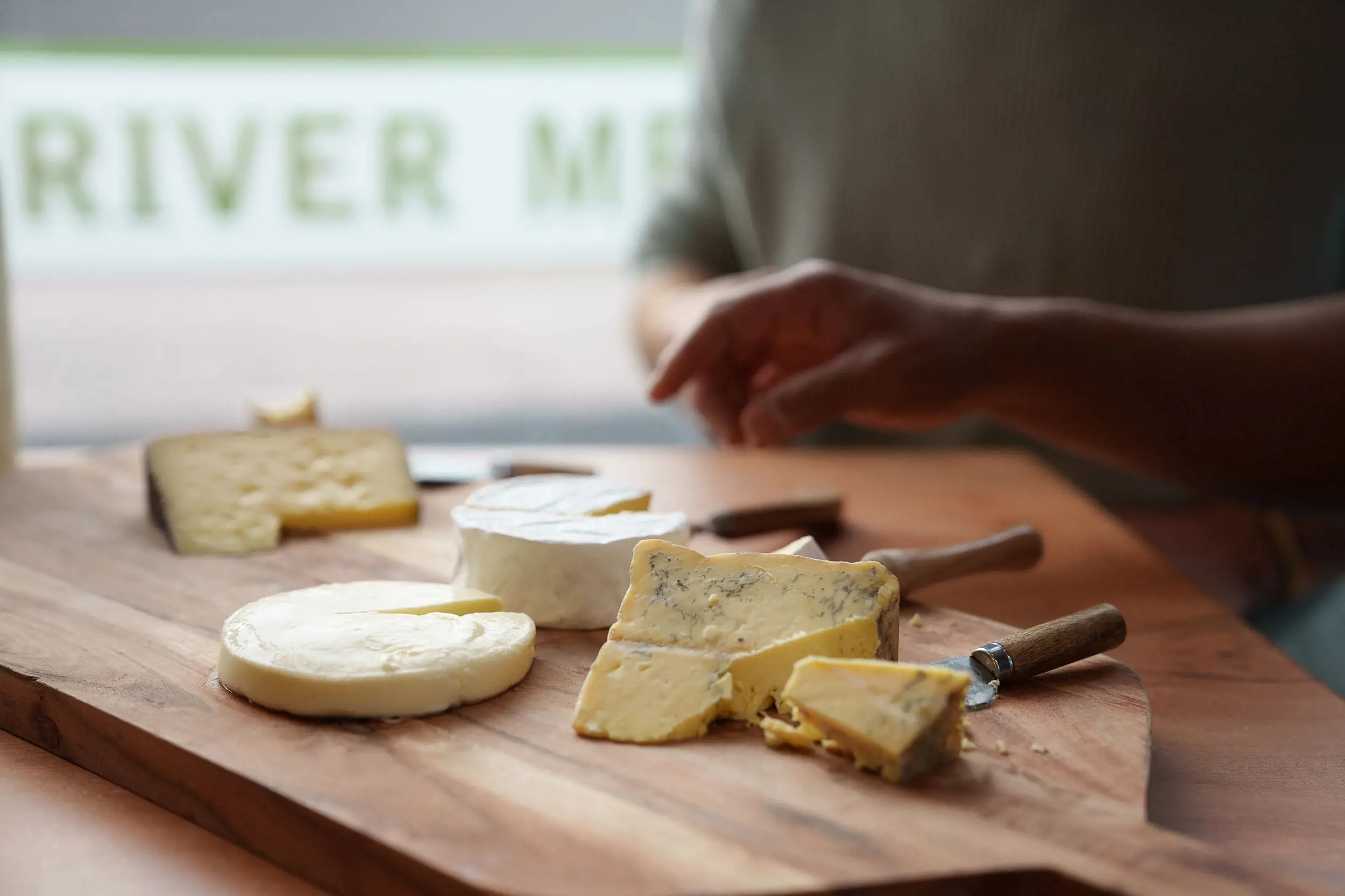 Cheeses are arranged on a wooden paddle. Chunks are cut from each cheese, with cheese knives lying beside them.