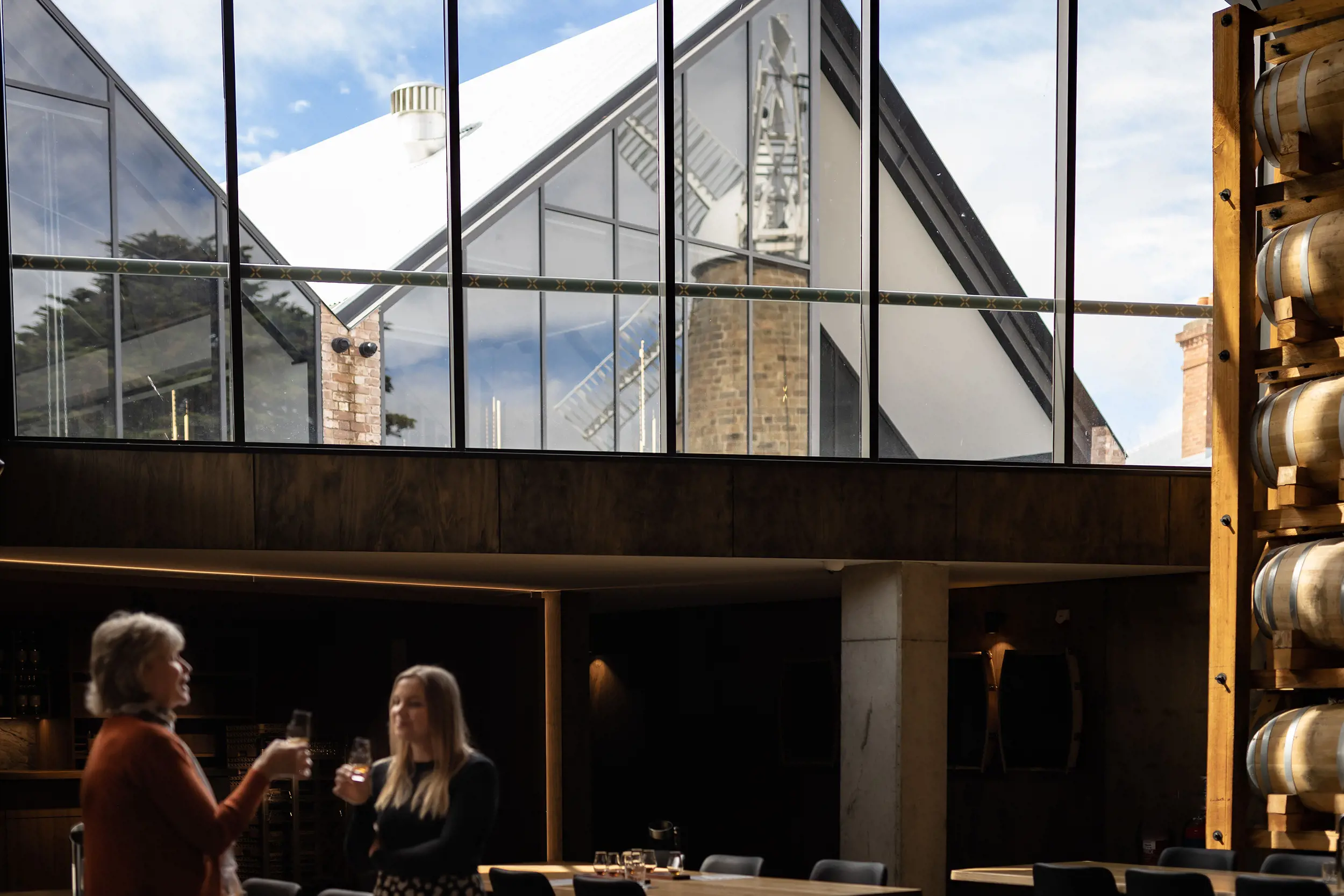 In a semi-underground cellar, two women are tasting whisky. A large window above them reveals the architecture outside, the glass reflecting an old windmill.