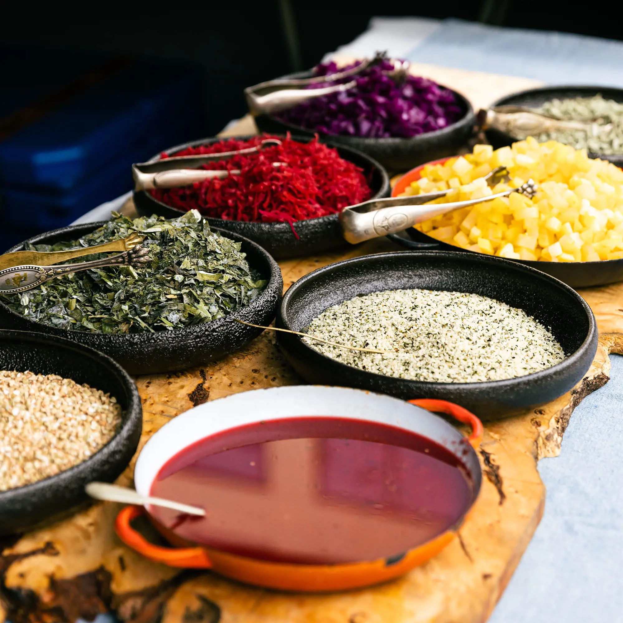 Eight wide flat bowls with a colourful range of ingredients in them, with silver tongs and metal spoons for serving.