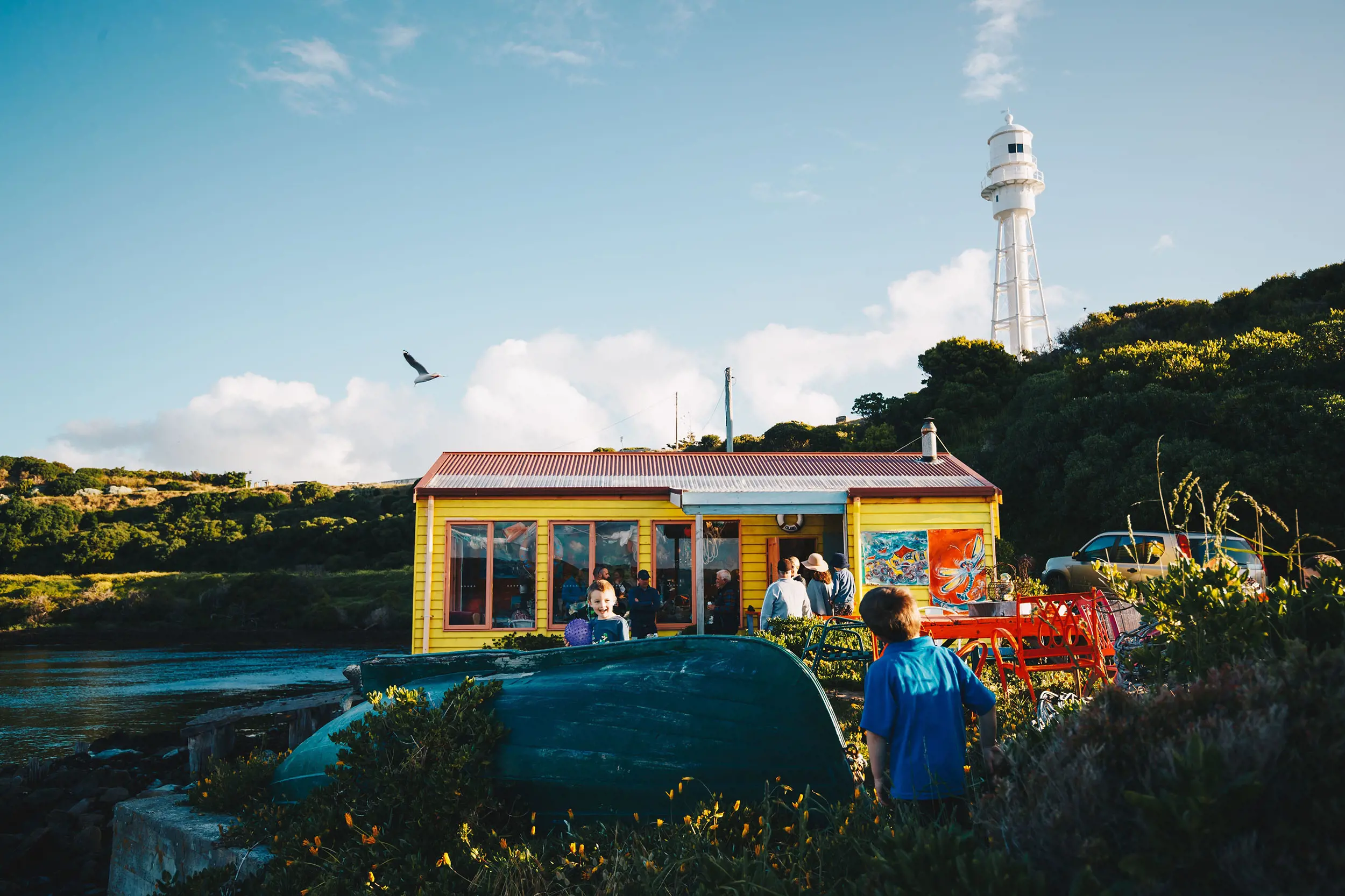 A small yellow weatherboard shack is nestled among the scrub by a beach. Children play next to an upturned dinghy out the front, and a white lighthouse structure rises in the distance.