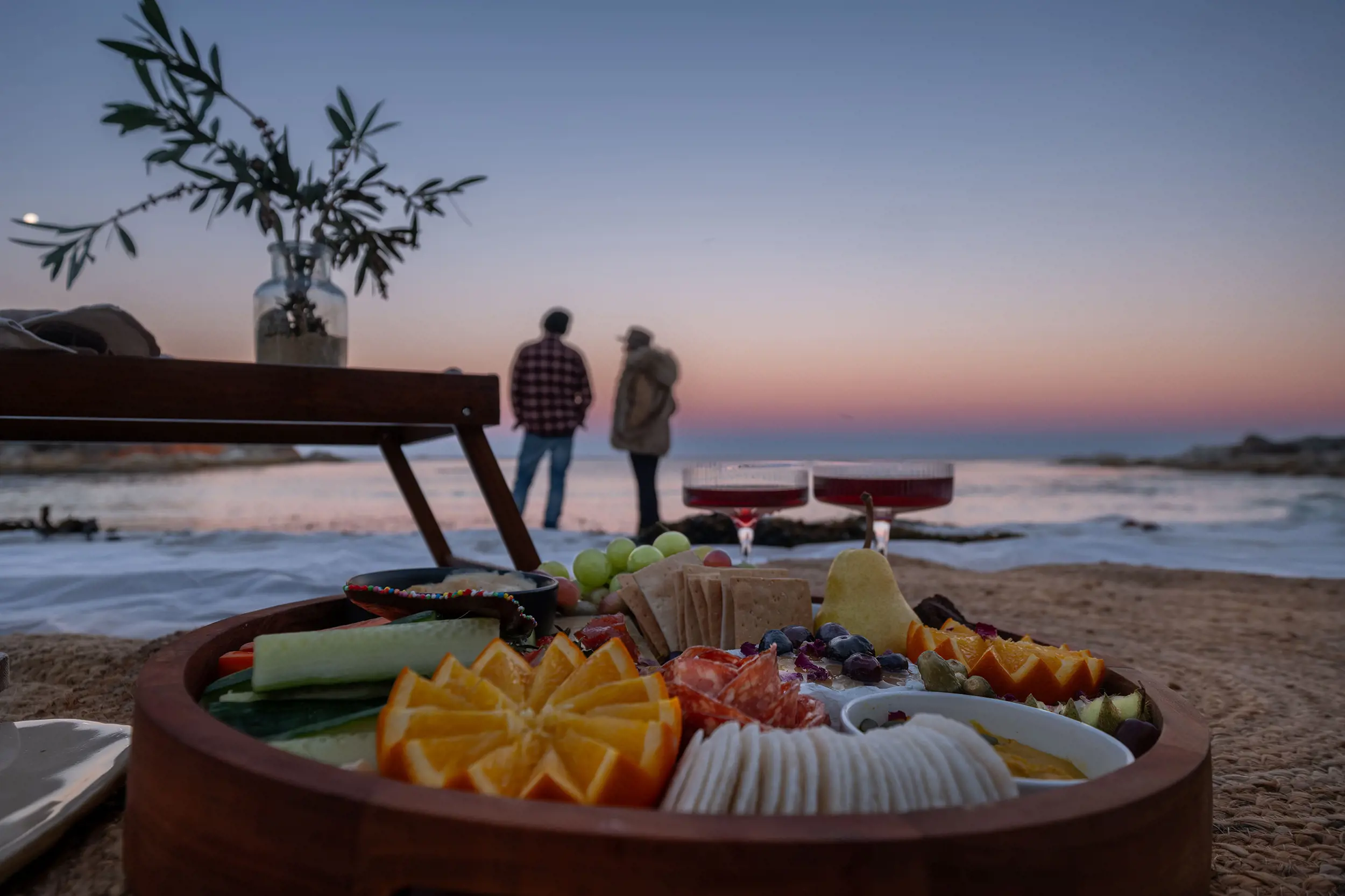 A close-up of a plate of cut fruits, cheeses and crackers, and two drinks sit on the sand at a beach. In the background stand two people with their feet in the water, looking at the sunset.