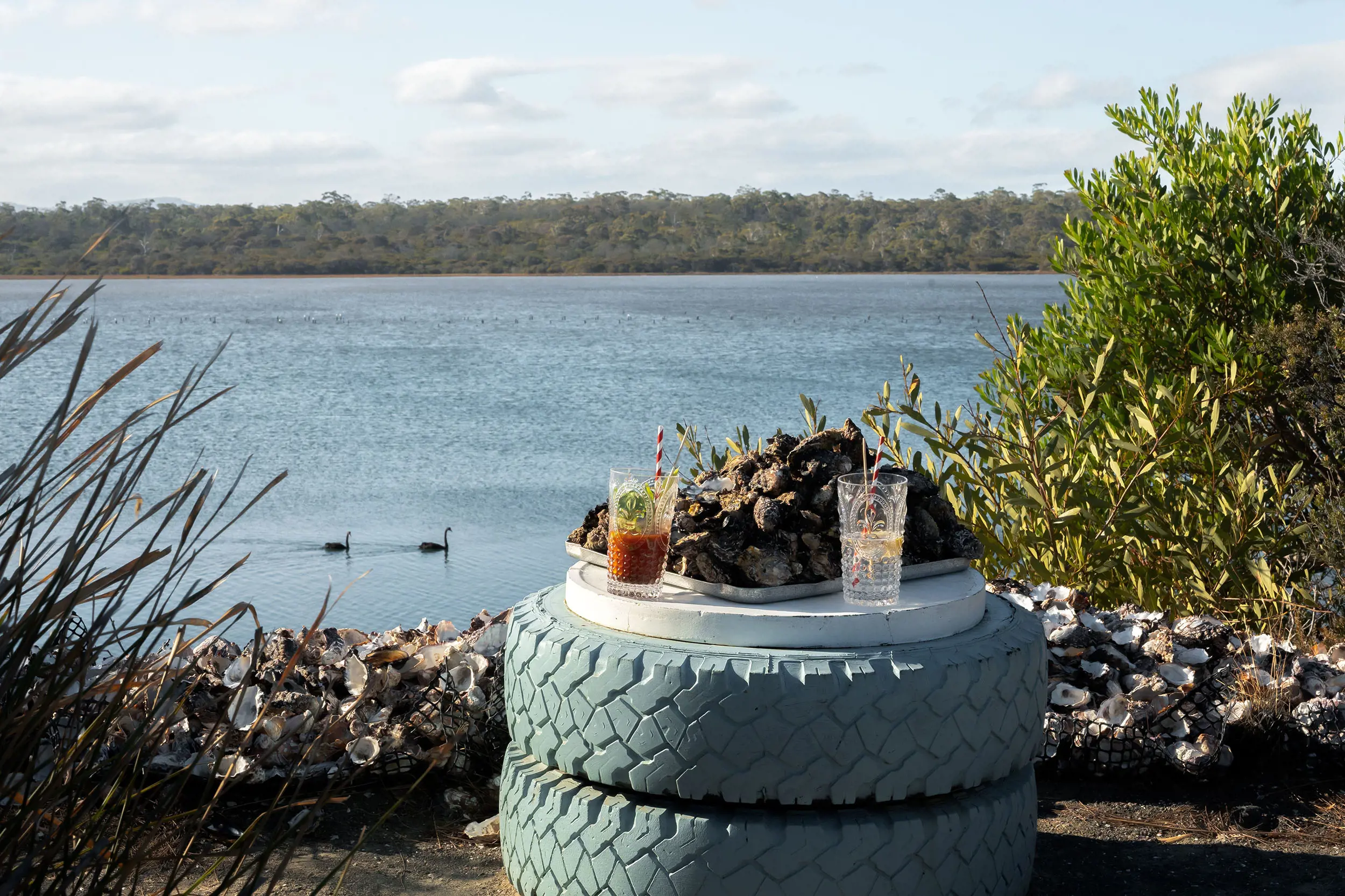 A rustic table made of two painted tyres carries a tray piled high with unshucked oysters and two drinks, next to a picturesque river view.