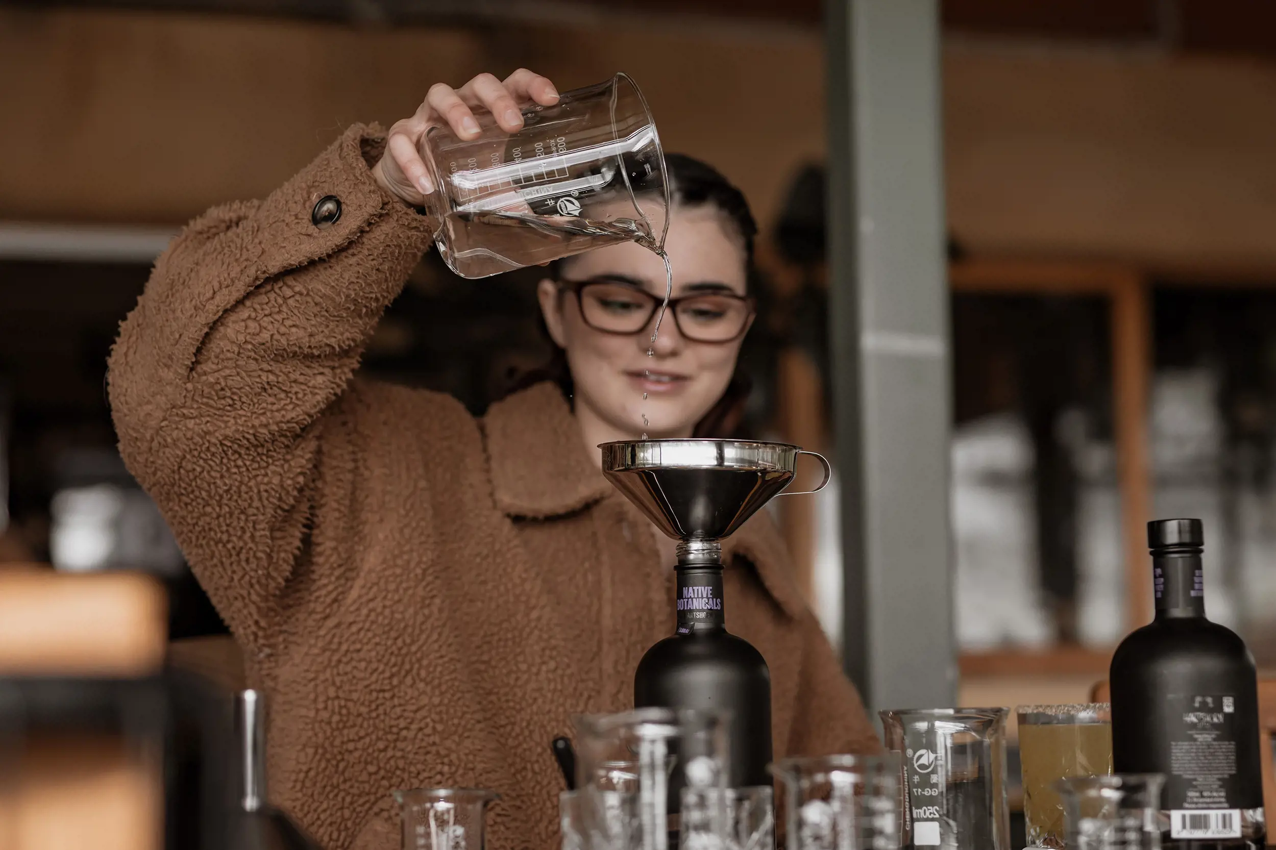 A woman slowly pours a clear liquid from a glass beaker into a bottle with a silver funnel. Other bottles and glassware sit in front of her.