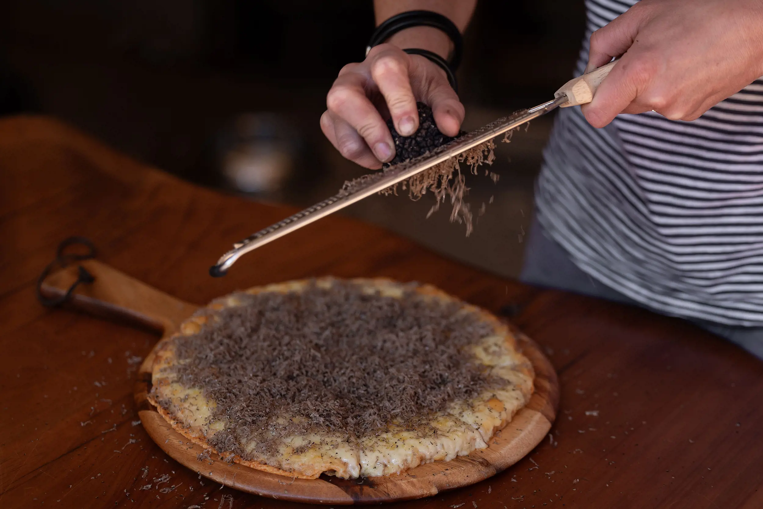 A person holds a microplane over a fresh pizza on a wooden paddle, grating a whole truffle onto the pizza.