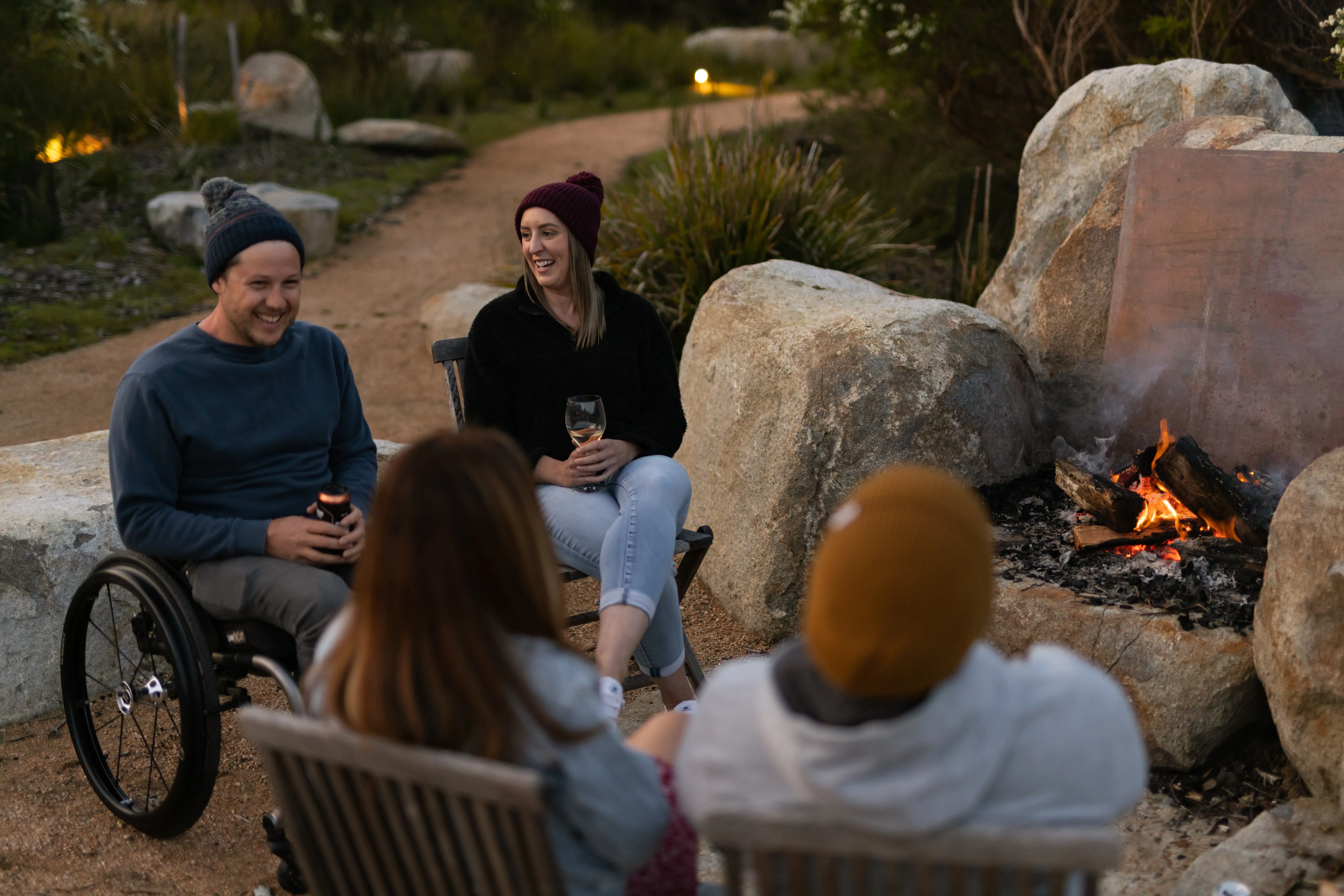 Four people sit around a campfire, three in wooden outdoor chairs and one in a wheelchair. They are holding drinks and wearing warm outdoor clothing and beanies.