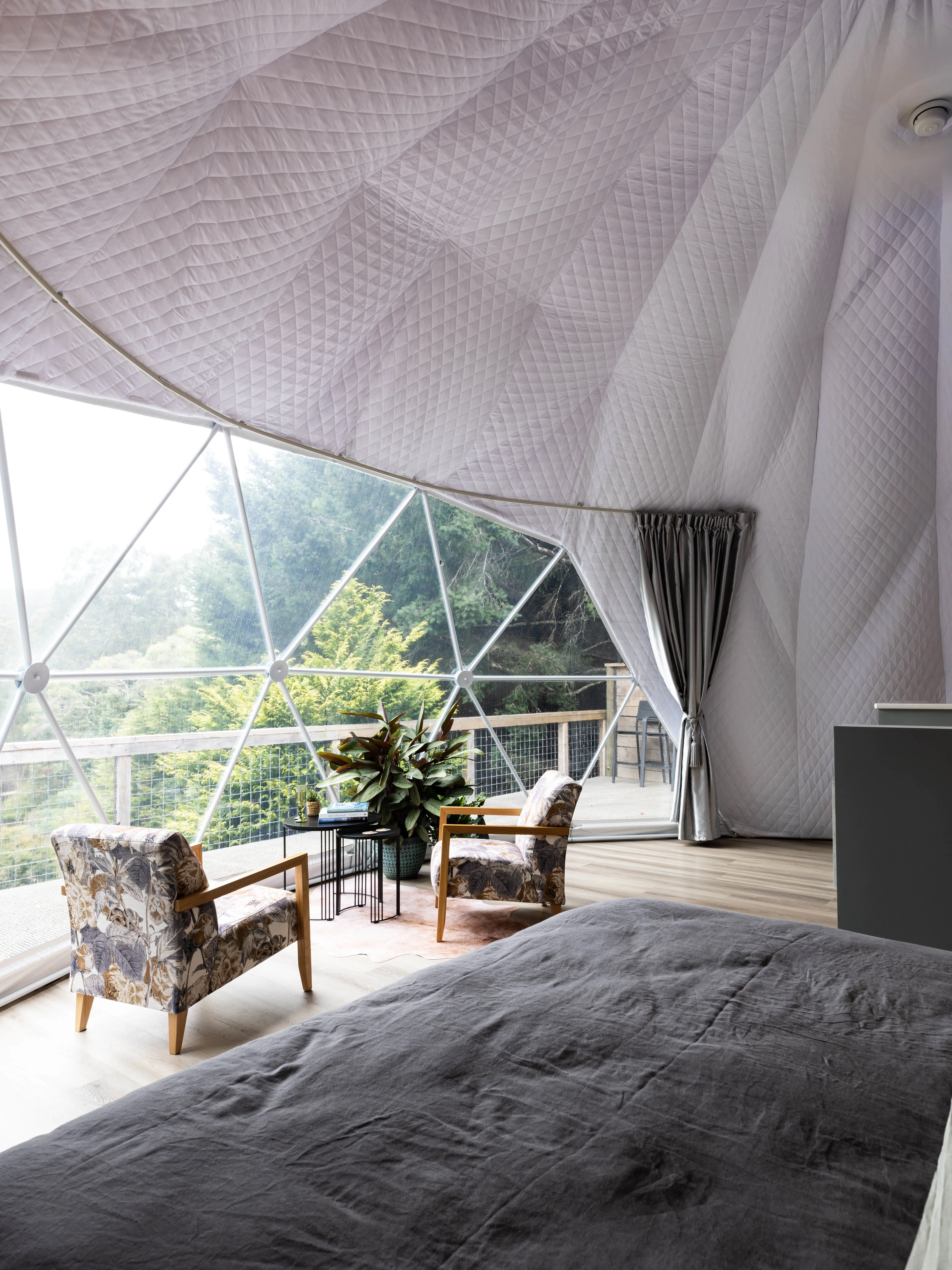 Inside a large plush dome structure with several clear triangular panels forming a window out to the bush. A table and armchairs sit by the window.