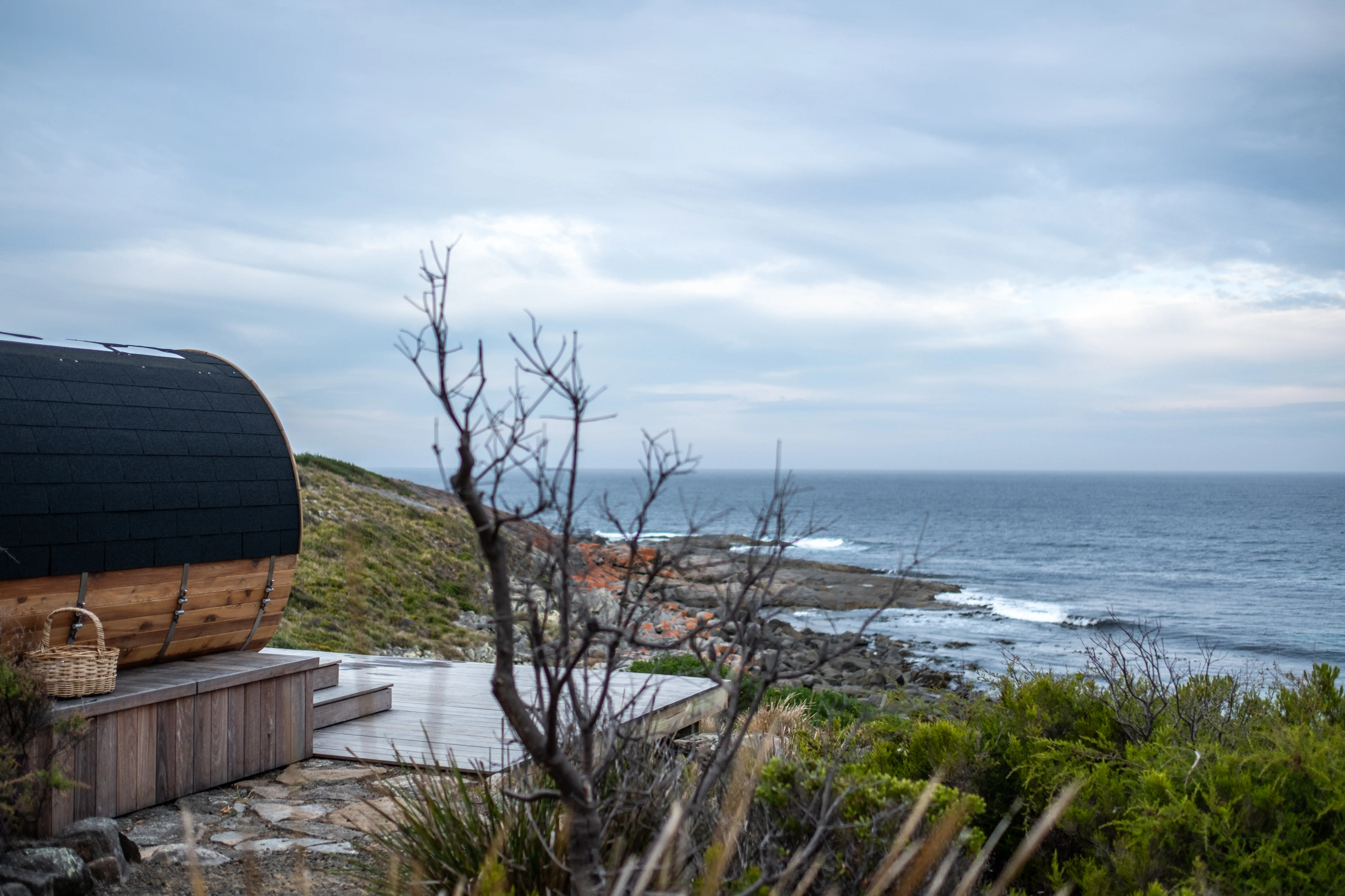 A wooden outdoor sauna sits among the bush overlooking the ocean. Waves are crashing below and the sky is full of dark clouds.