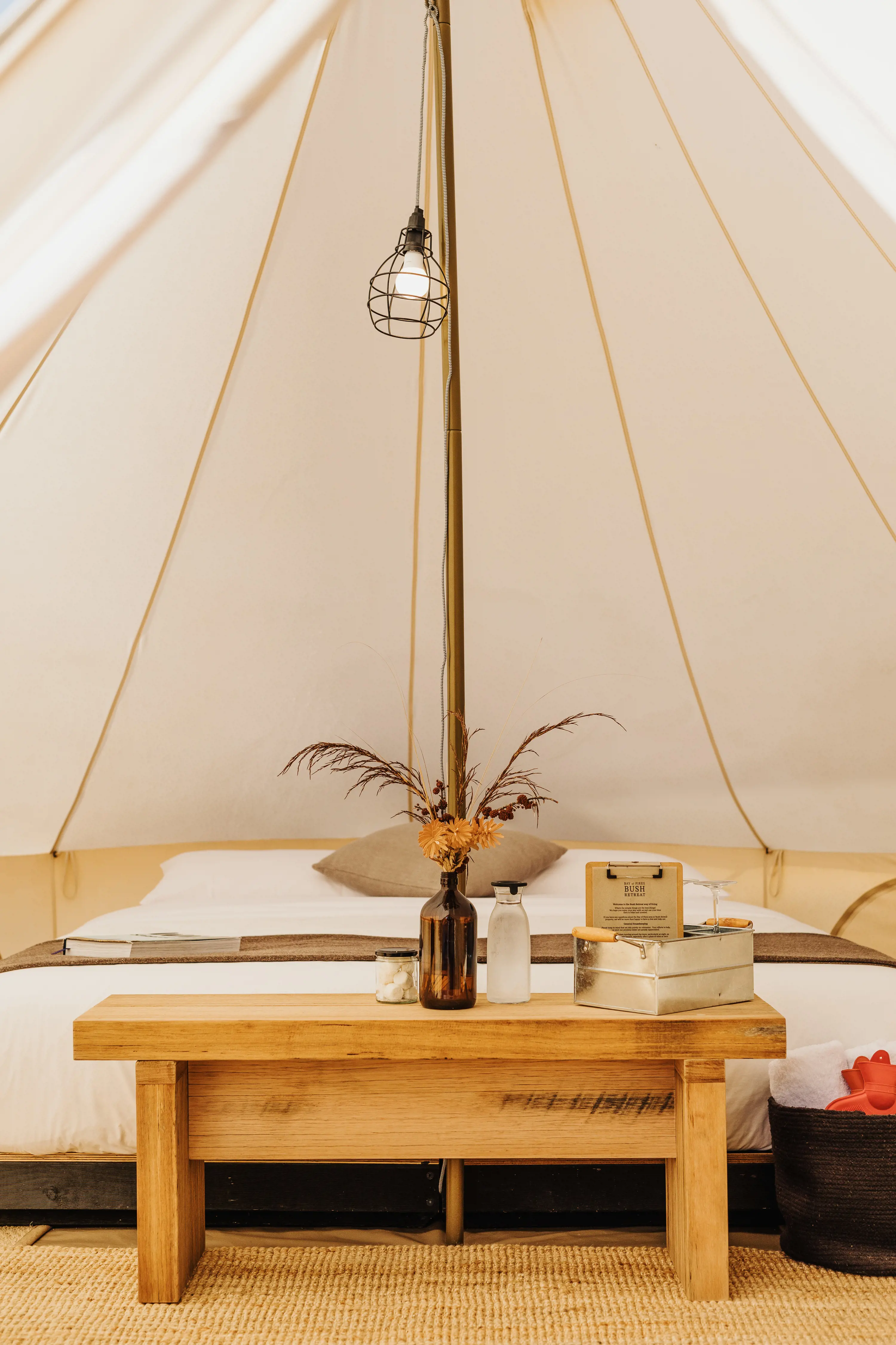 Inside a light canvas bell tent, a lamp hangs from the central post and underneath sits a bed, wooden end table and amenities.