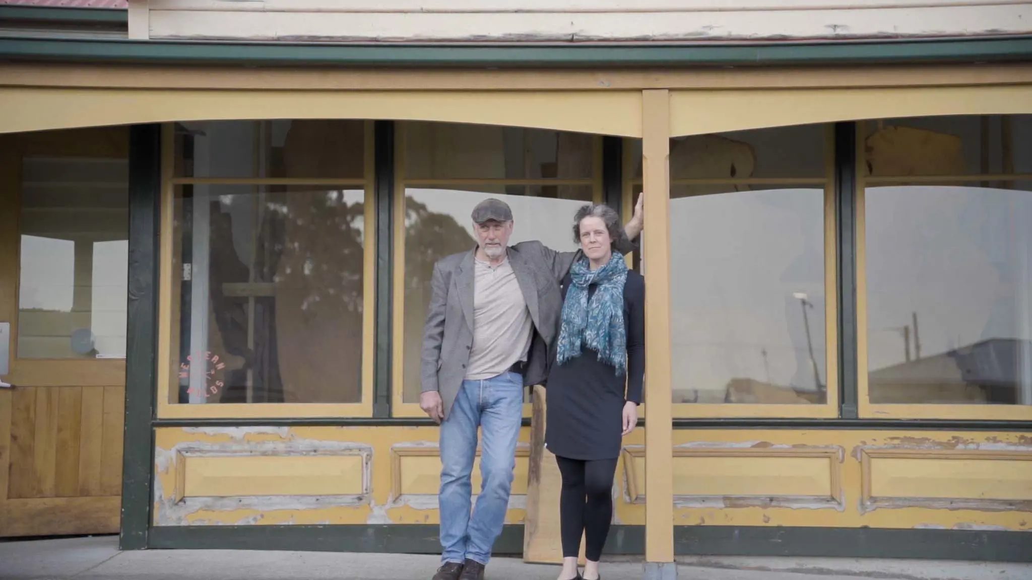 A man wearing a flat cap and jacket and a woman with a large scarf stand under a verandah in front of a shop with large glass windows. 