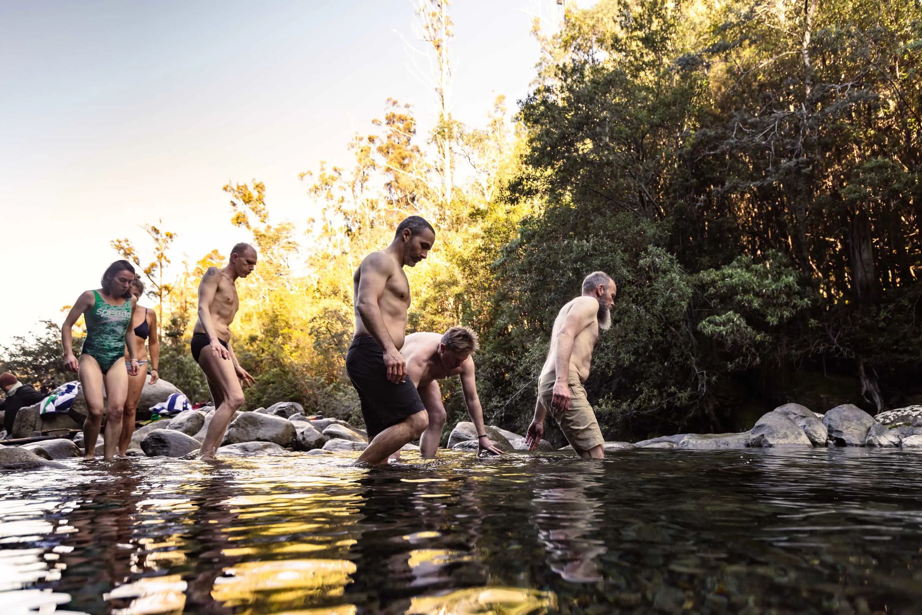 A group of people in swimming gear walk from the rocky edges of a pond in the forest into deeper water. 