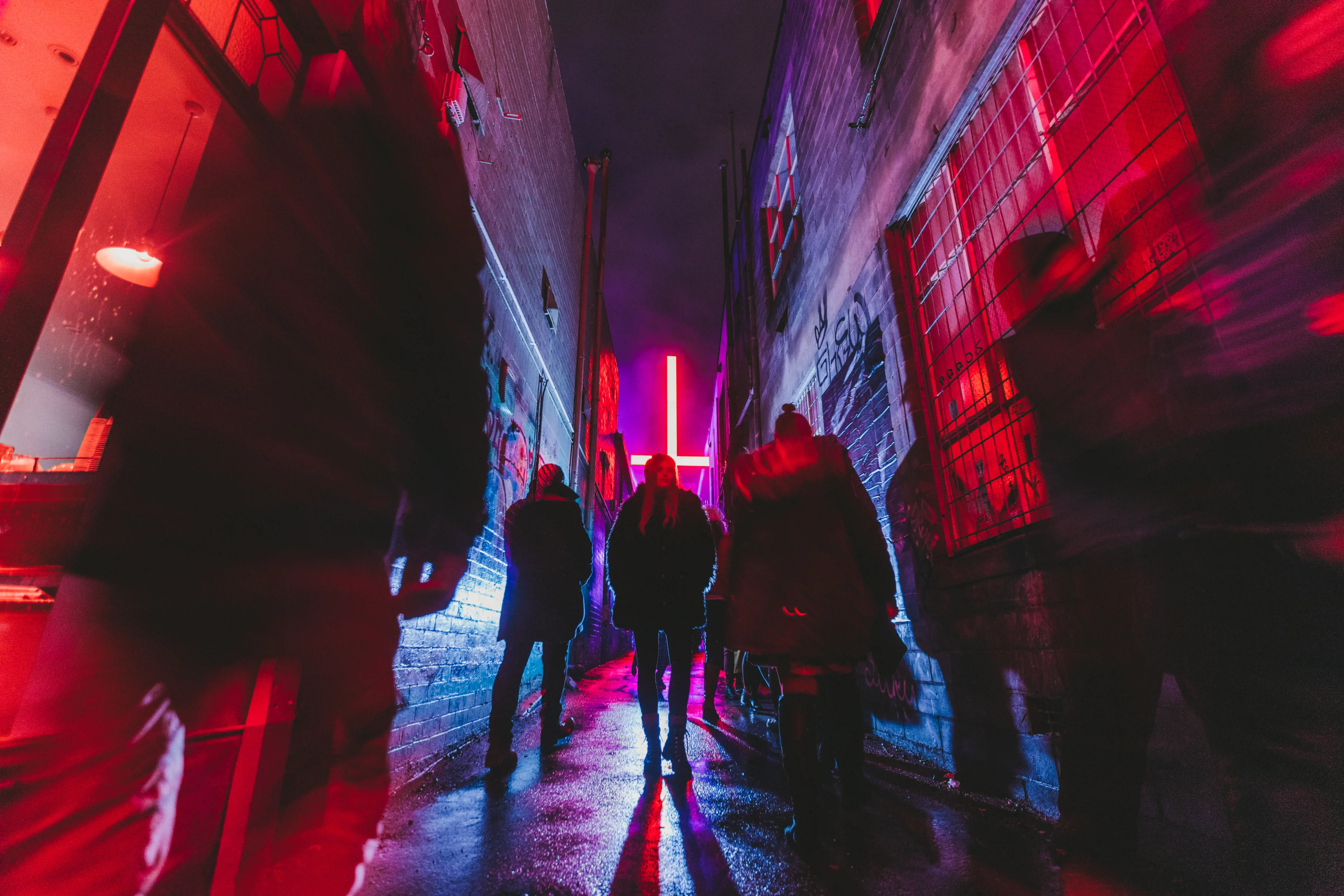 A large neon cross is suspended above people walking down a narrow alleyway at night.